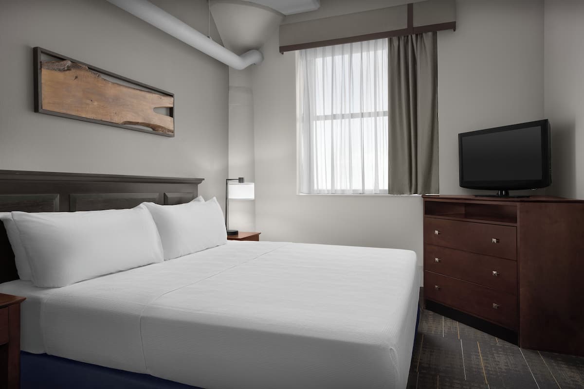 A hotel room with a large bed, a flat-screen TV, a wooden headboard, a window with curtains, and a wooden dresser.