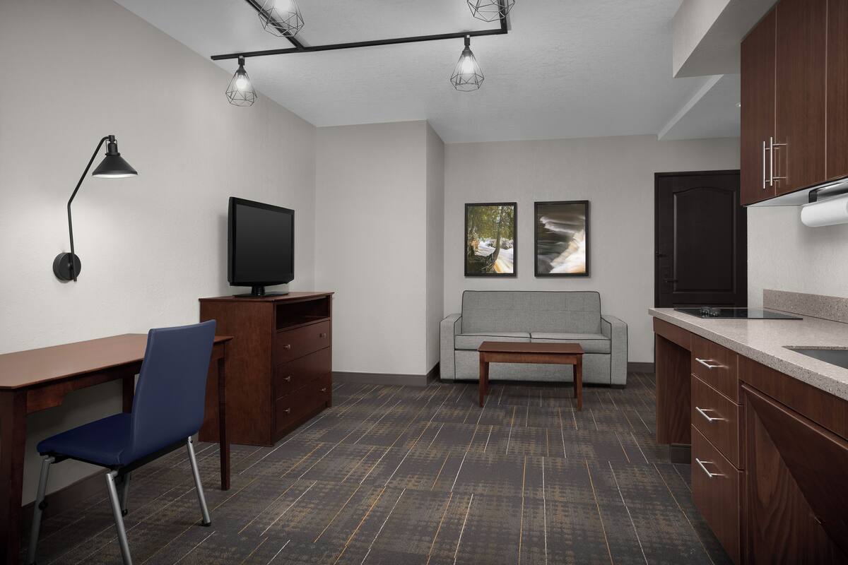 A modern kitchen with a built-in oven, a dining area with a wooden table and blue chair, a gray sofa, and a TV on a stand.