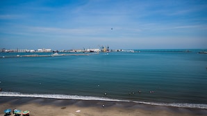 Vista costera con playa arenosa, mar tranquilo y estructuras industriales a lo lejos.