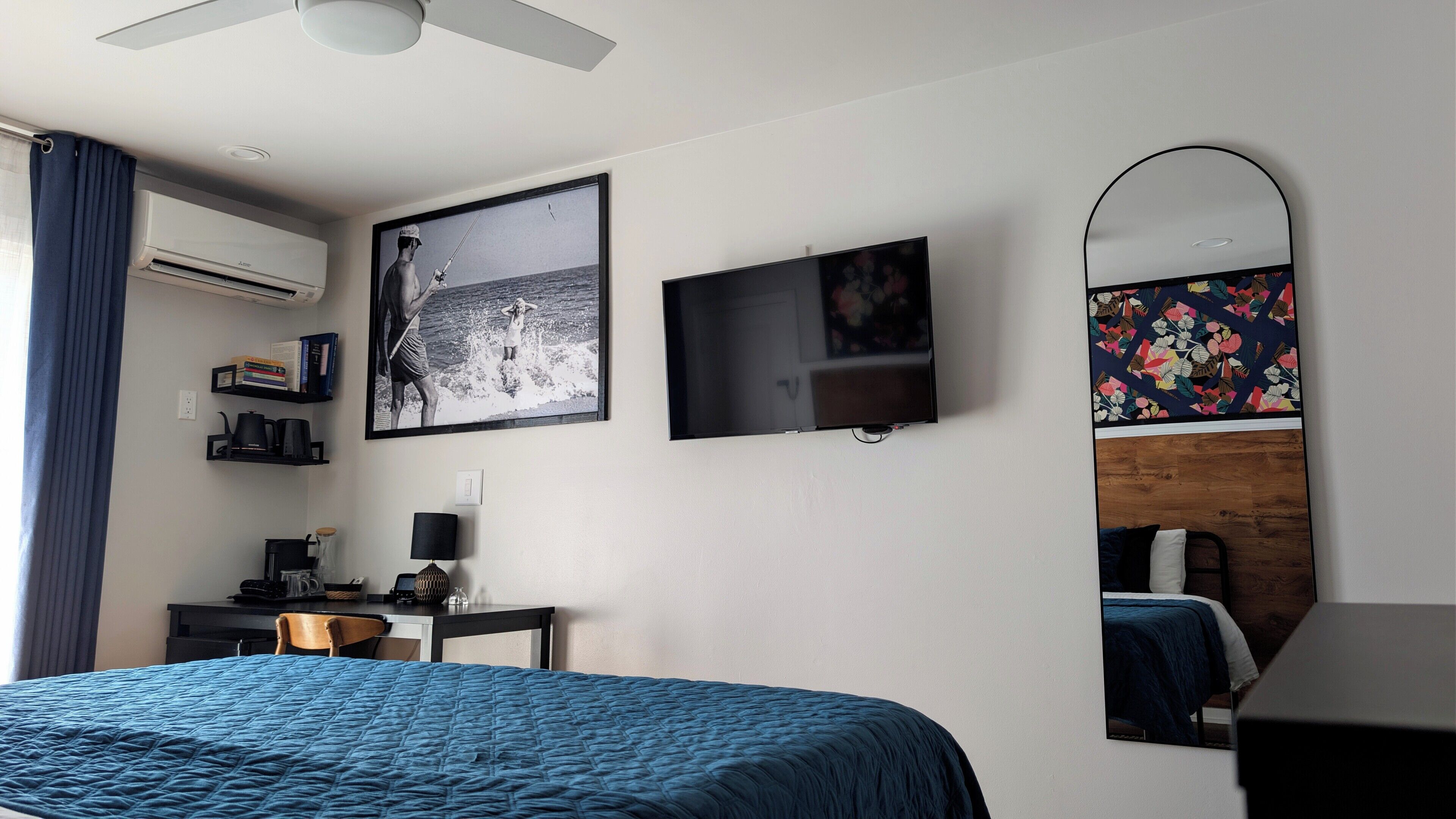 A bedroom with a bed, a desk, a chair, a TV, a mirror, and an air conditioning unit.