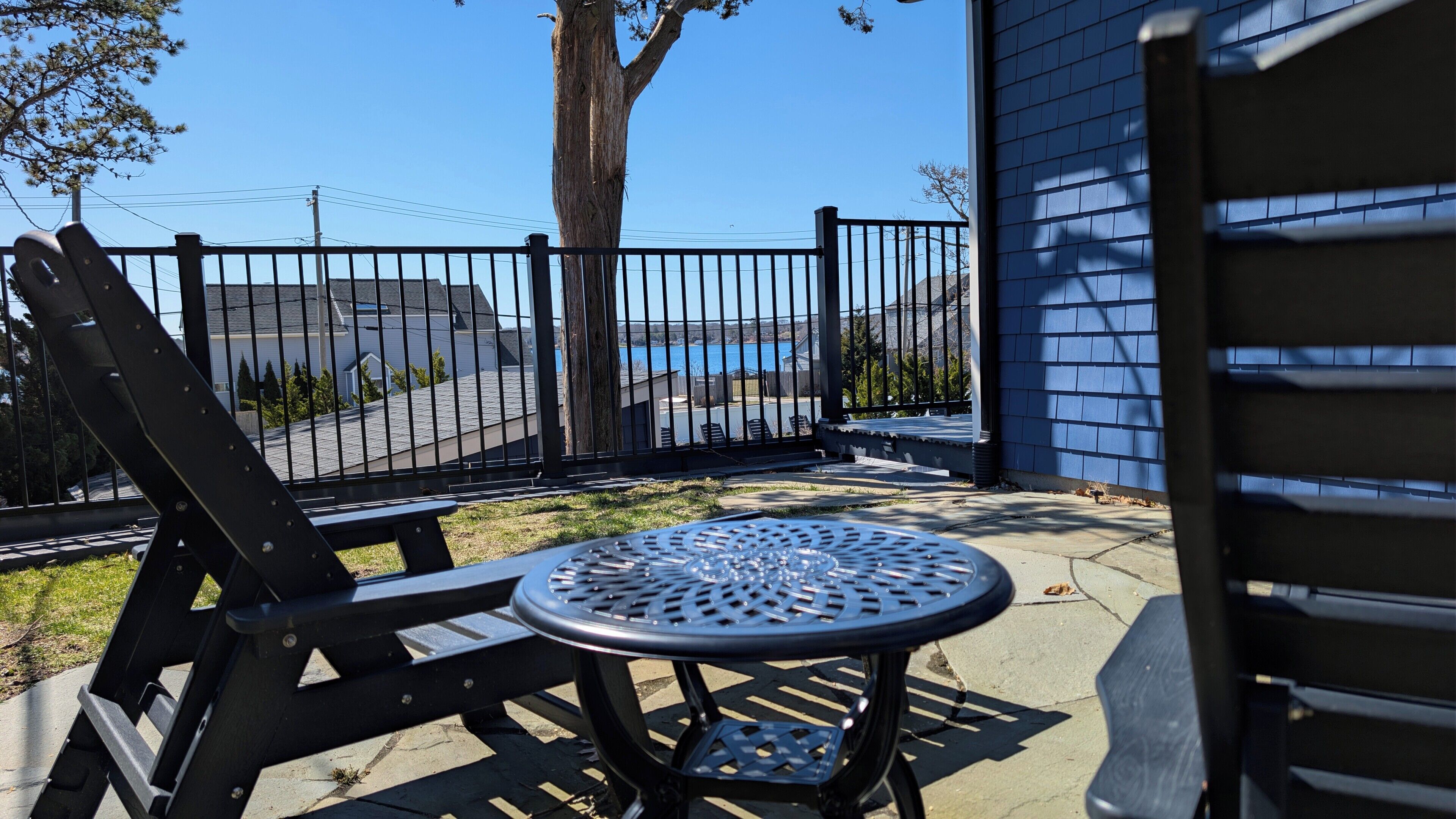 Un patio con una mesa y sillas de metal, vista hacia un cuerpo de agua y un edificio azul al fondo.