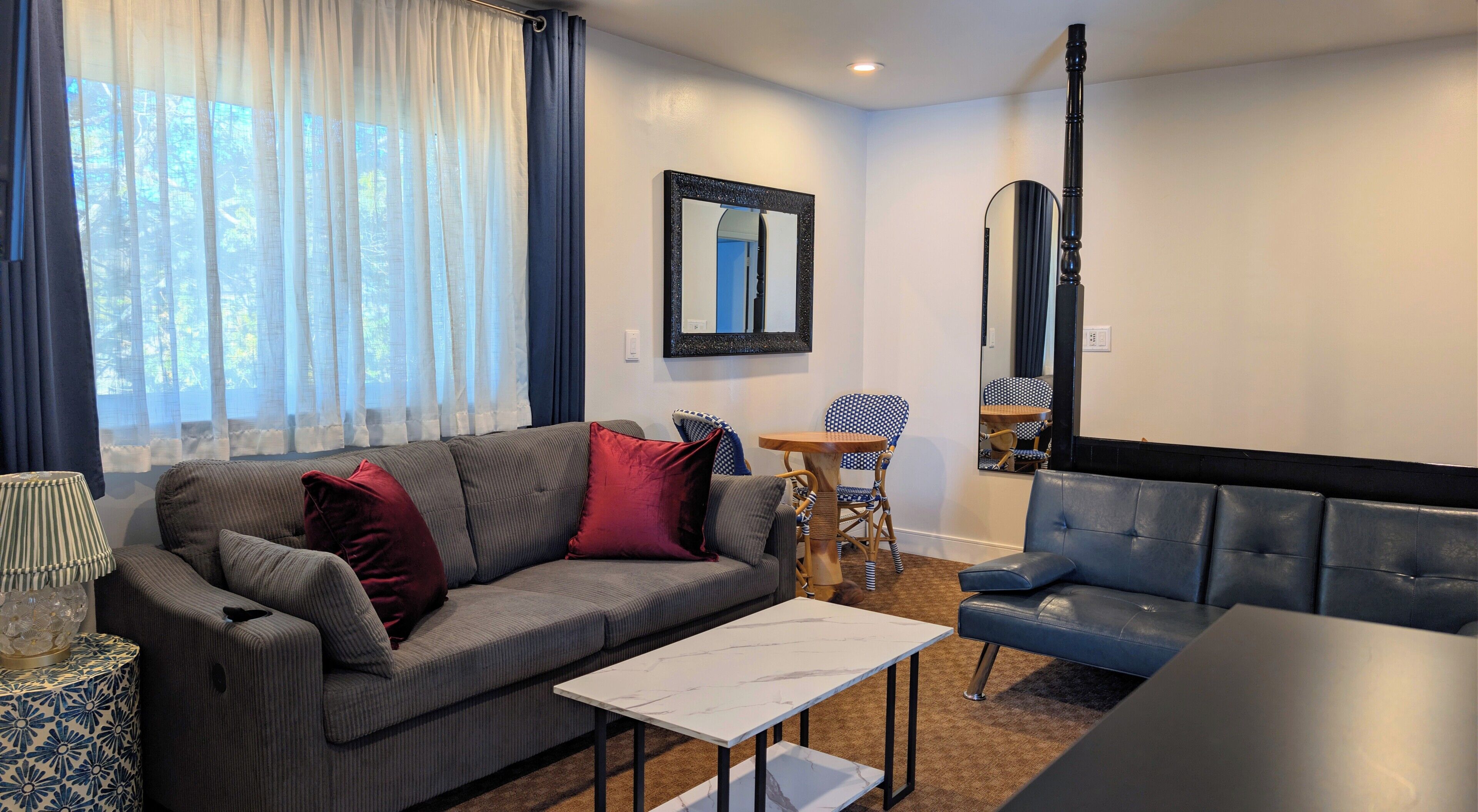 A living room with a sofa, a coffee table, a dining table, and chairs. There is a window with curtains, a mirror on the wall, and a lamp on a side table.