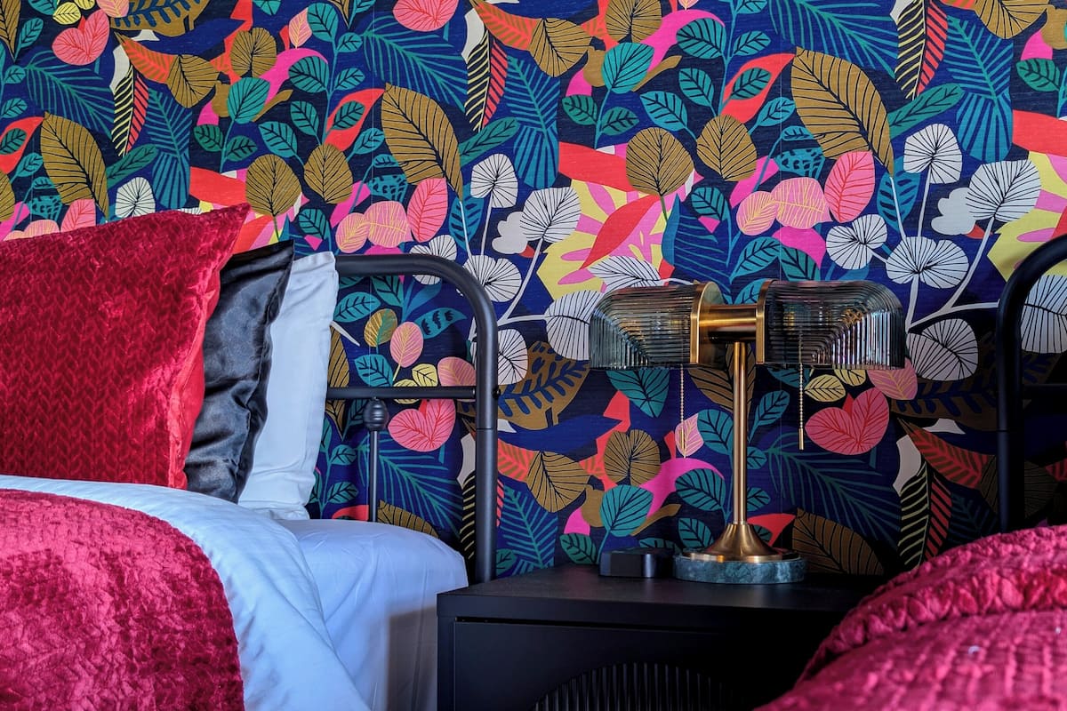 Una cama con ropa de cama roja, una mesita de noche negra con una lámpara, y una pared con diseño de hojas.