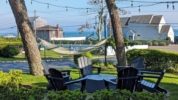 An outdoor seating area with Adirondack chairs and a table, surrounded by American flags and flowers, with a view of houses and a body of water in the background. - Ocean View Terrace Inn (Hampton Bays)