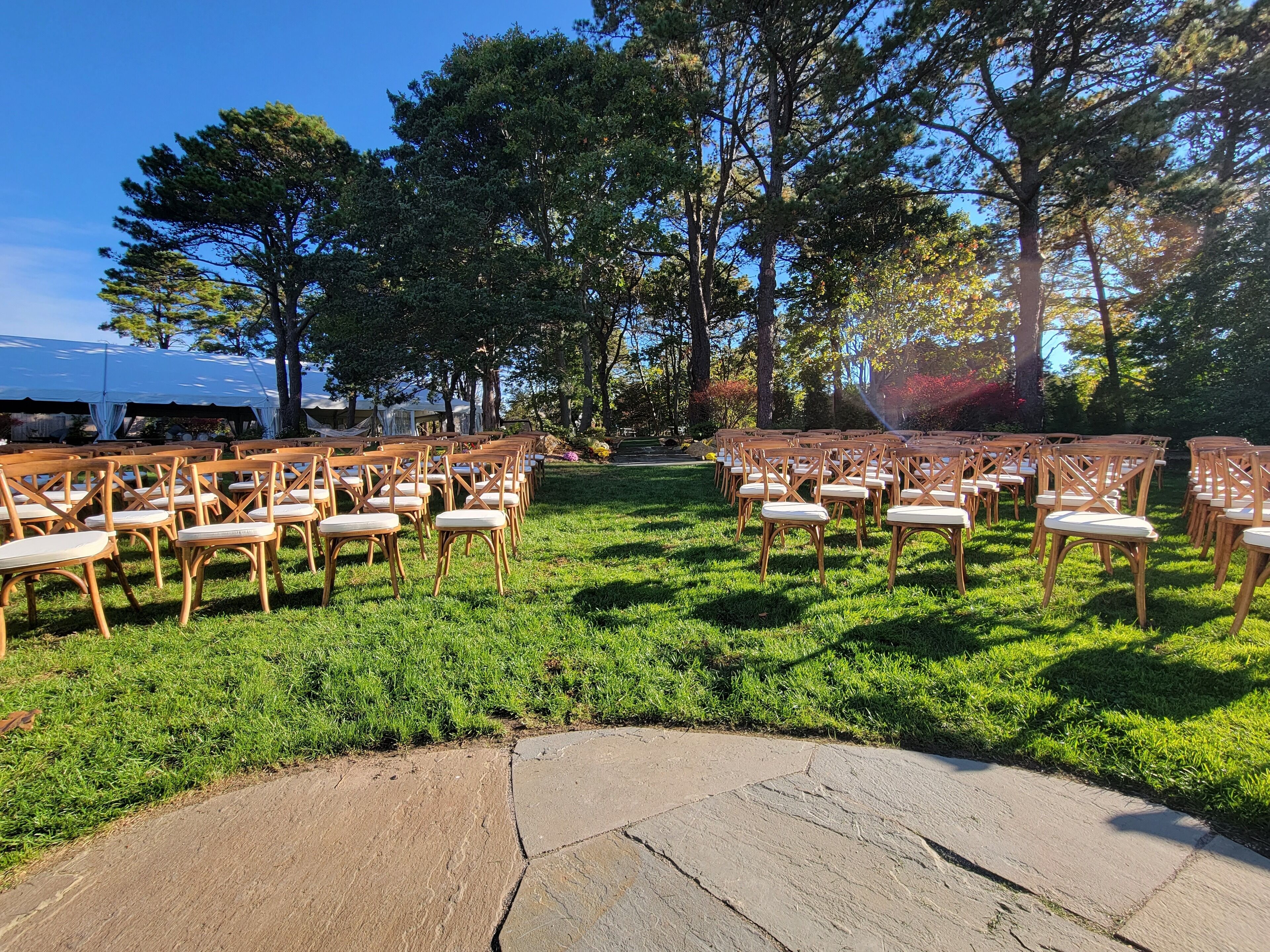 An outdoor seating area with wooden chairs and tables, surrounded by trees and a grassy lawn.