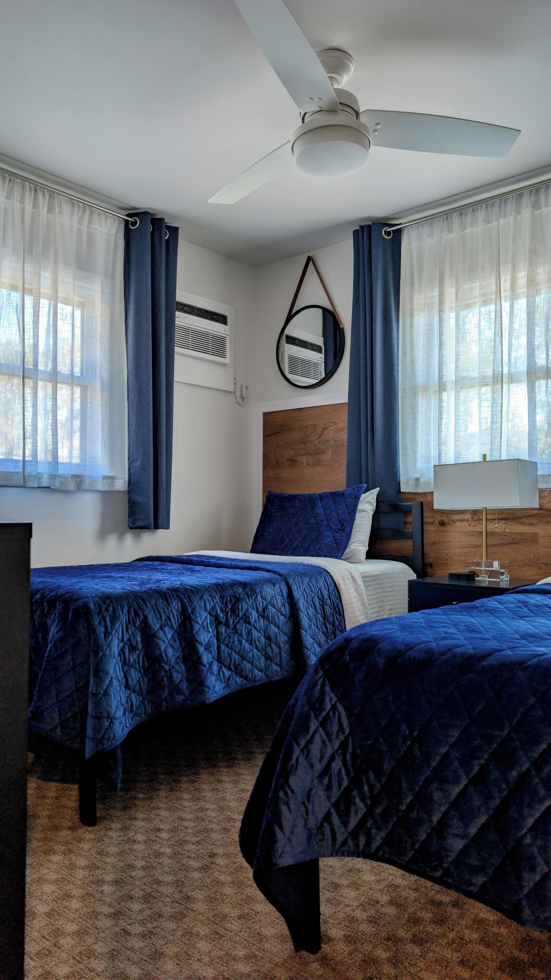 Une chambre d’hôtel avec deux lits, un ventilateur de plafond, la climatisation, une tête de lit en bois, une table de chevet avec une lampe et des rideaux légers.