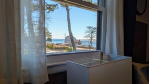 A window with a view of a building and trees, a small table with a lamp, and a curtain partially covering the window. - Ocean View Terrace Inn (Hampton Bays)