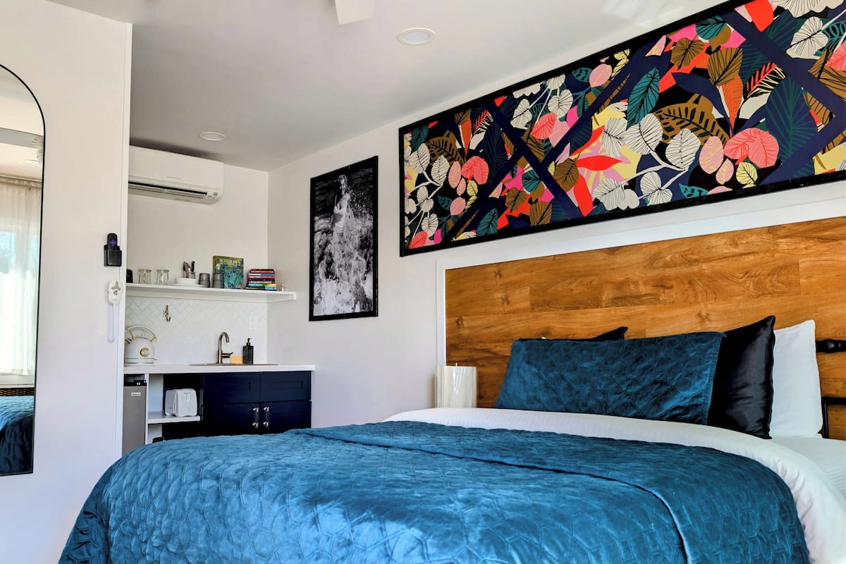 Un dormitorio con una cama grande, un cabecero de madera, un espejo, un aire acondicionado montado en la pared, un estante con libros y objetos decorativos, y un cuadro abstracto y colorido.