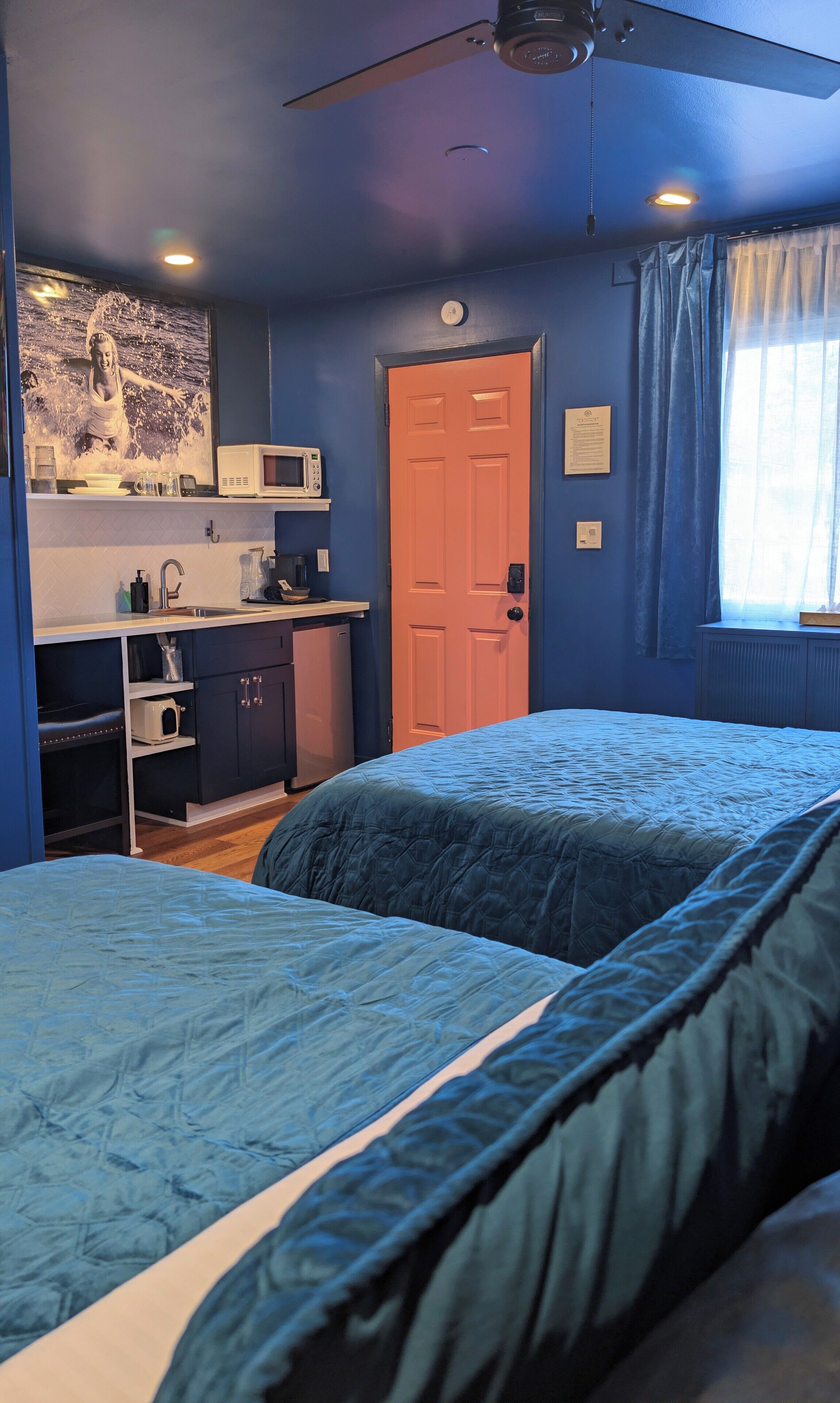 Une chambre avec deux lits, un coin cuisine, un ventilateur de plafond et une porte.