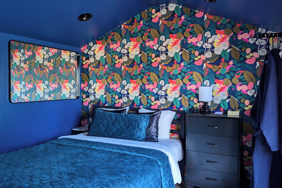 Un dormitorio con cabecera estampada de flores, cama con colcha azul, mesita de noche con lámpara y cómoda con cajones.