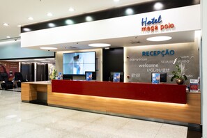 A hotel reception desk with a modern design, featuring a red and wooden color scheme and multilingual welcome signage. - Hotel Mega Polo (São Paulo)