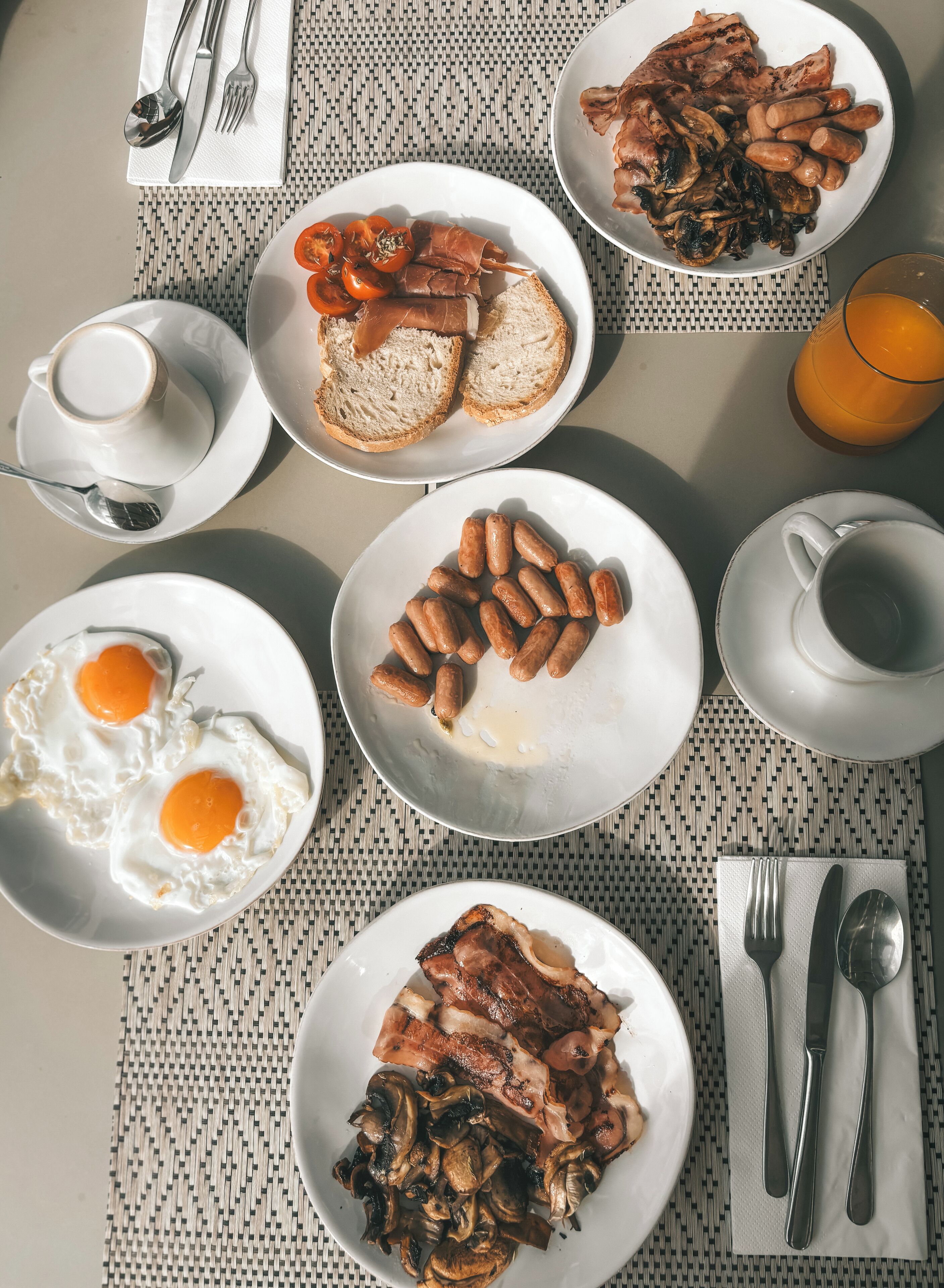 A breakfast setting with plates of eggs, sausages, bacon, and mushrooms, along with bread and a cup of coffee.
