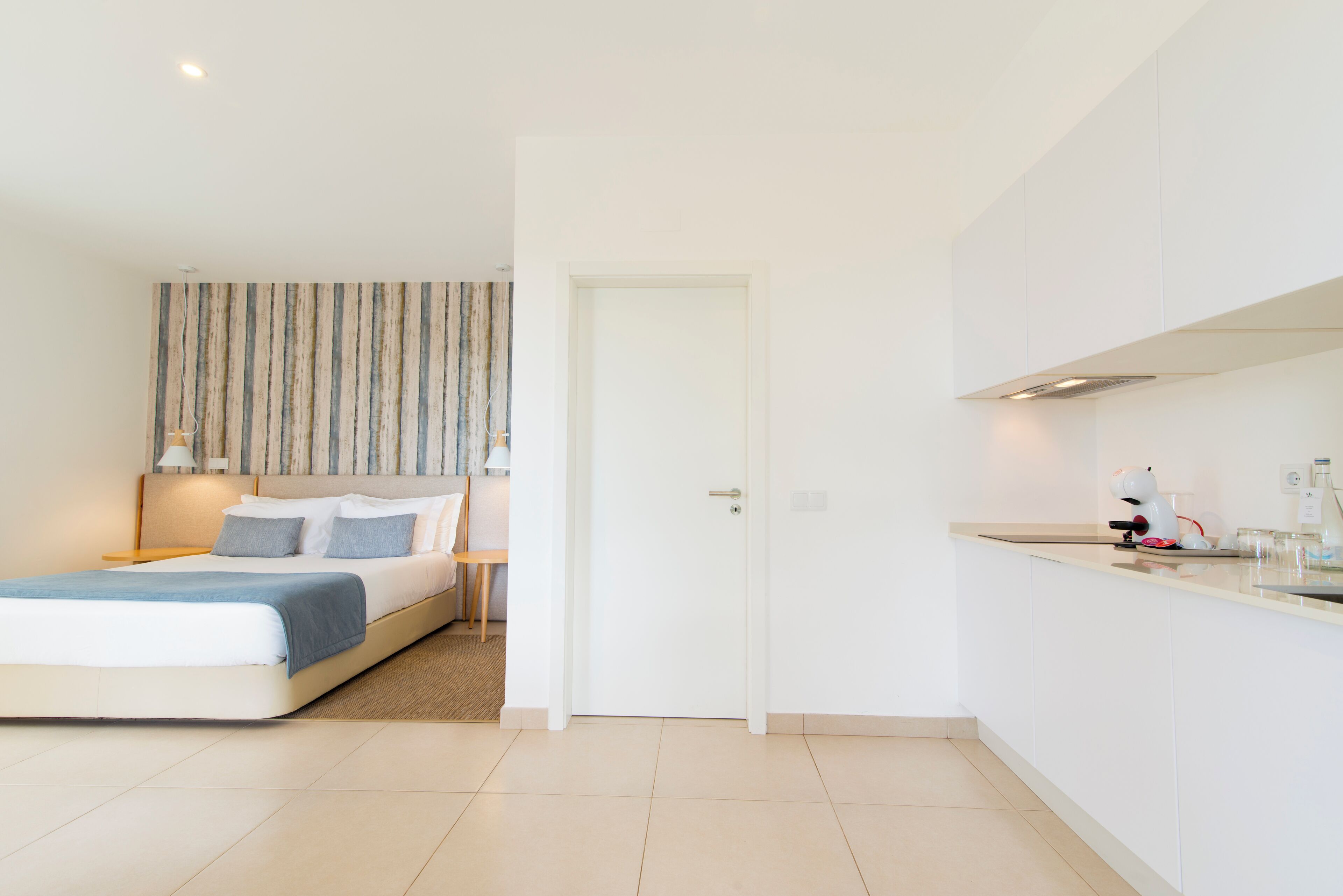 Ein modernes, minimalistisches Schlafzimmer mit einem großen Bett, einem Nachttisch und einem Beistelltisch. Eine Tür führt zu einem Küchenbereich mit weißen Schränken und einer Arbeitsplatte.