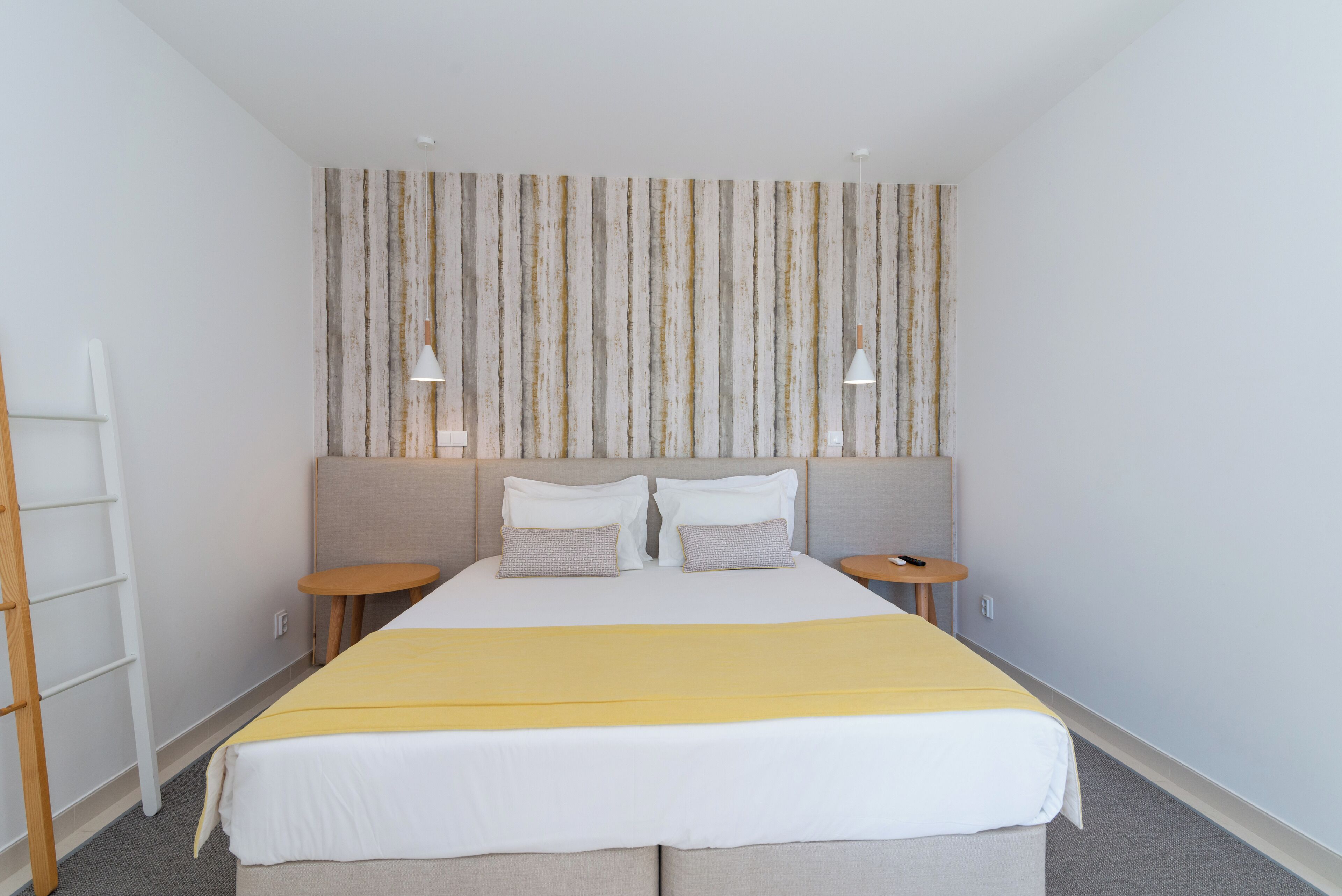 Habitación de hotel ordenada con una cama grande, dos mesitas de noche, una pequeña mesa redonda y una escalera apoyada en la pared.