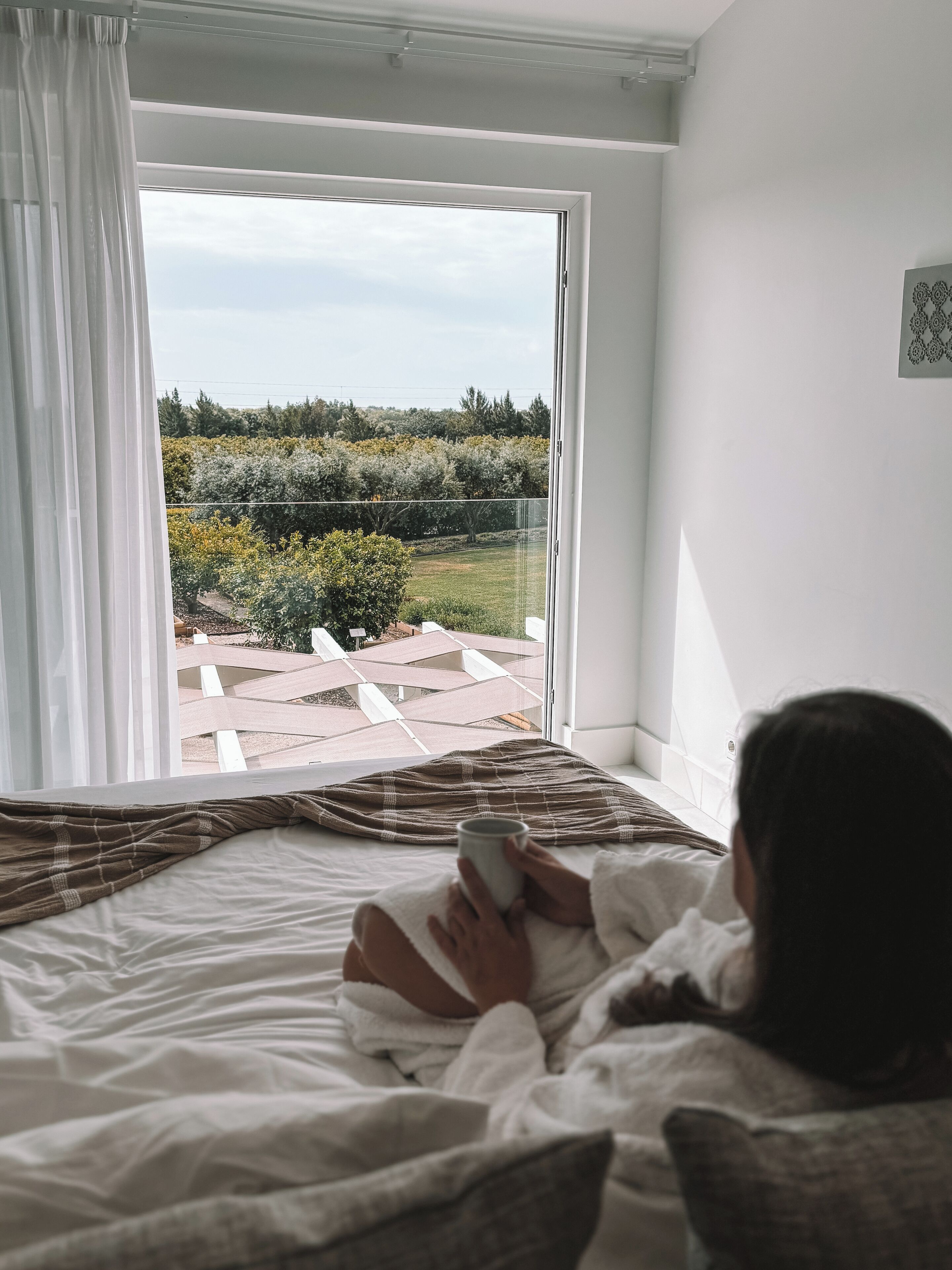 A bedroom with a large window, a bed, and a view of a garden and pathway.