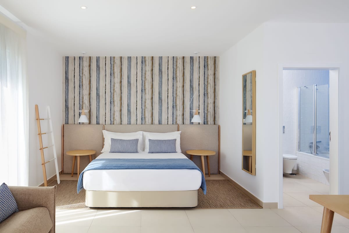 Une chambre d’hôtel moderne avec un grand lit, deux tables de chevet, une petite table ronde, un canapé et une vue sur la ville par la fenêtre.