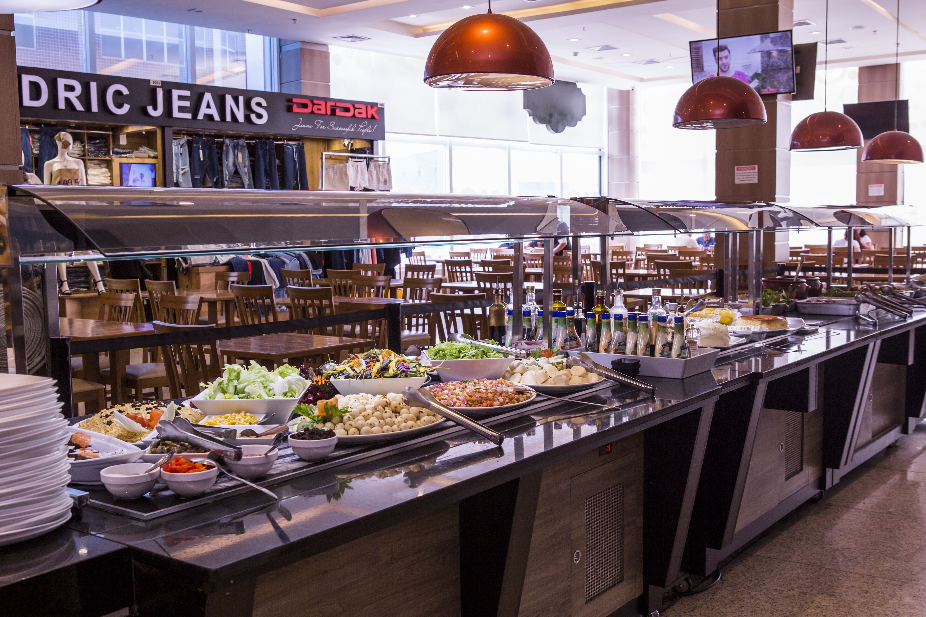 Un espace buffet proposant divers plats, un magasin de vêtements appelé « DRIC JEANS » en arrière-plan, et un coin salon avec des tables et des chaises.