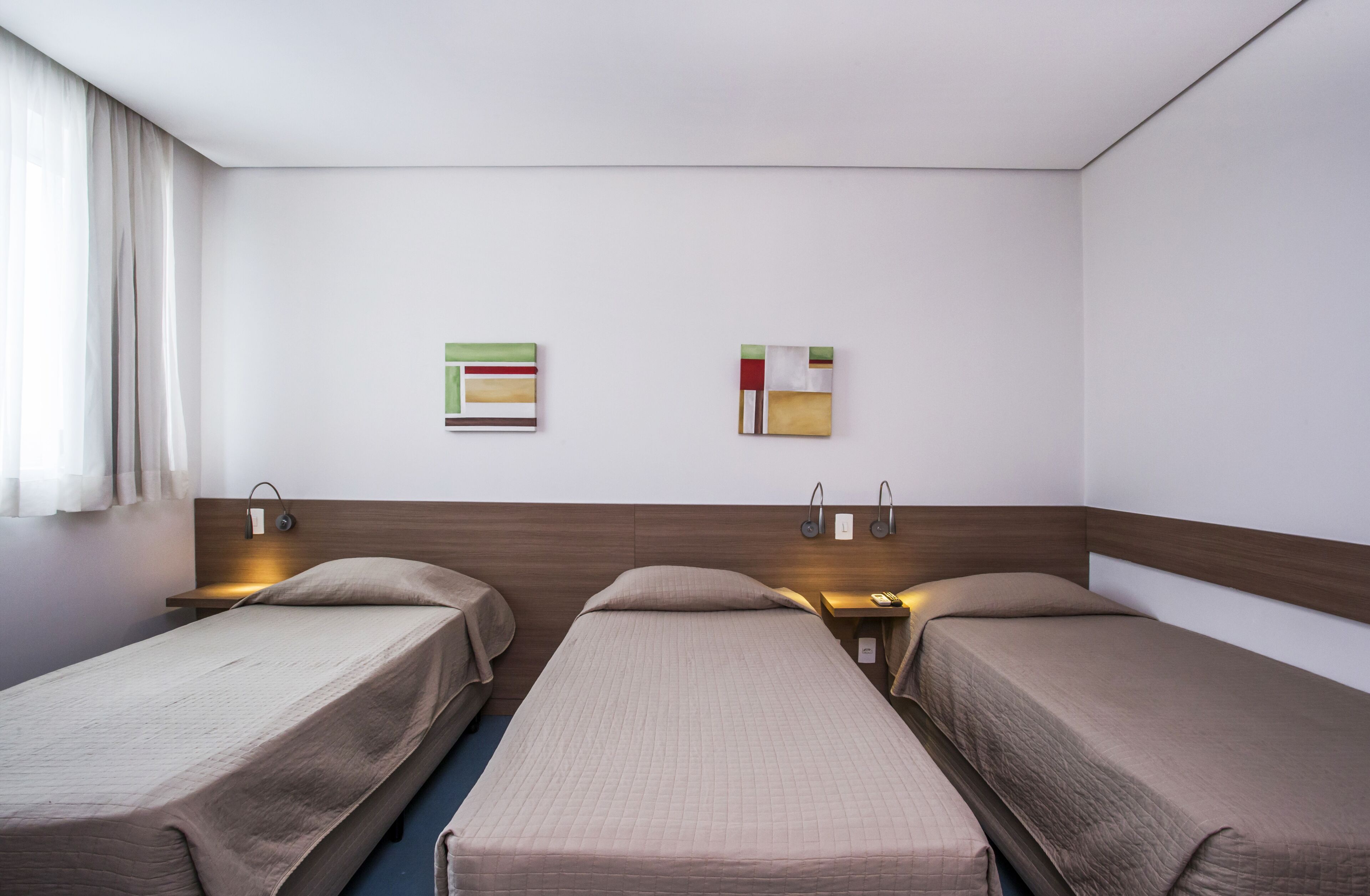 A hotel room with two single beds, a wooden headboard, bedside tables with lamps, and two abstract wall art pieces.
