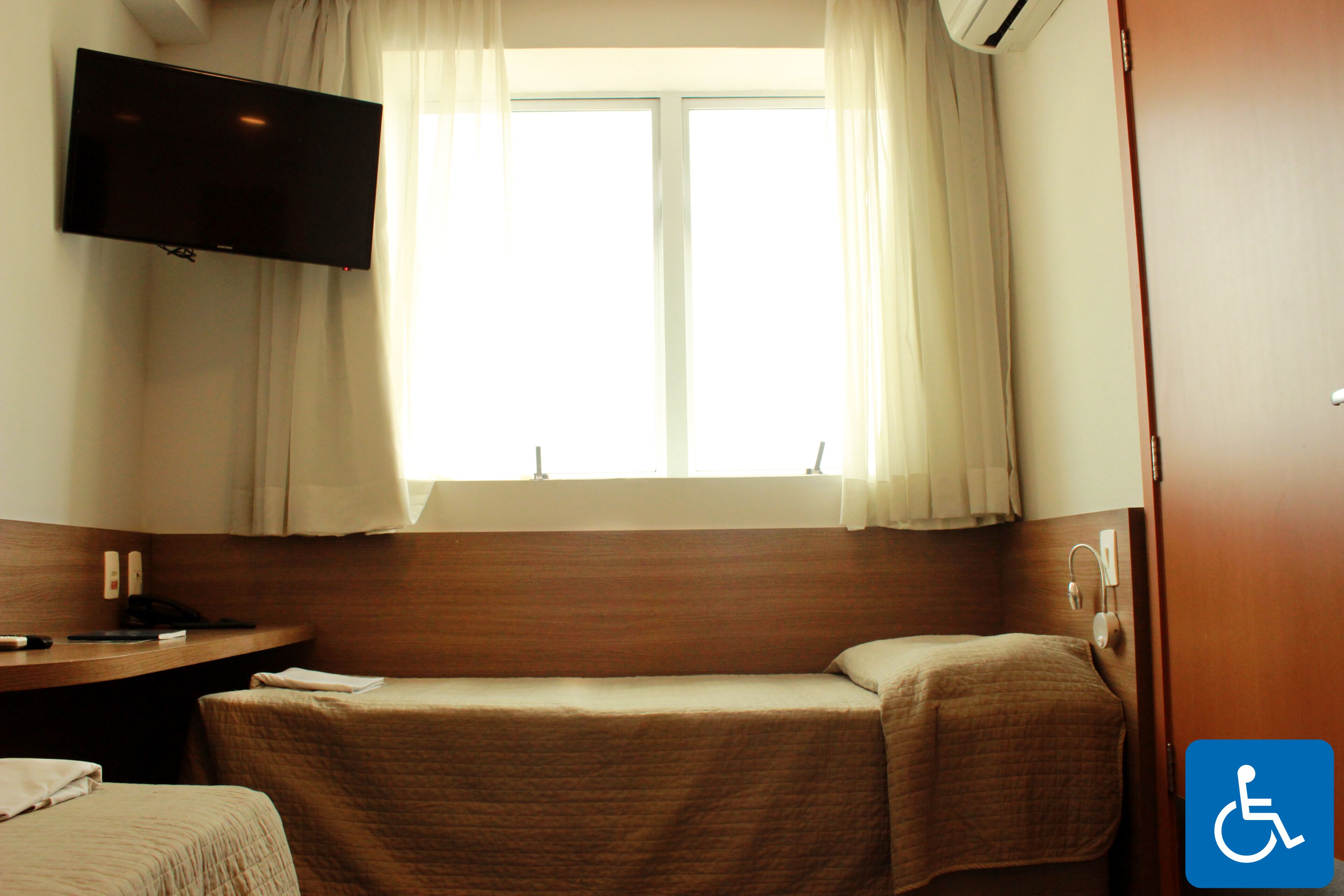 A hotel room with a bed, a bedside table, a TV mounted on the wall, and a window with curtains.