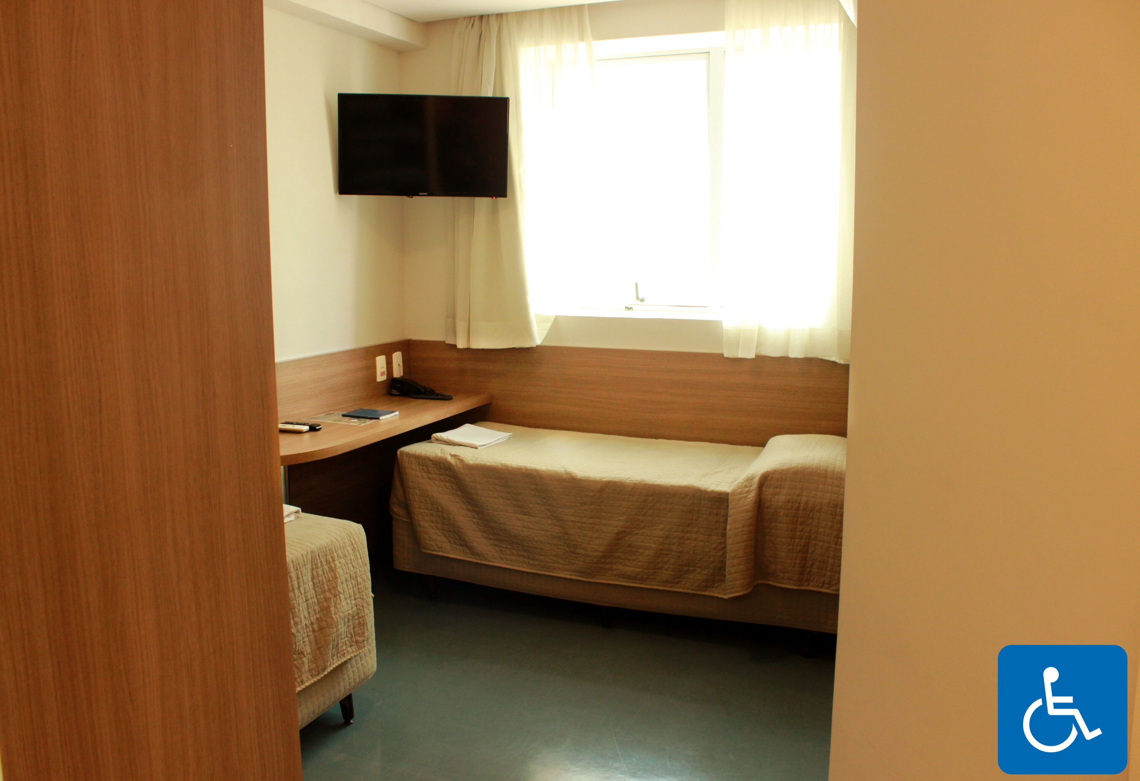 Ein kleines Hotelzimmer mit einem Bett, einem Schreibtisch, einem Stuhl, einem wandmontierten Flachbildfernseher und einem Fenster mit Vorhängen.