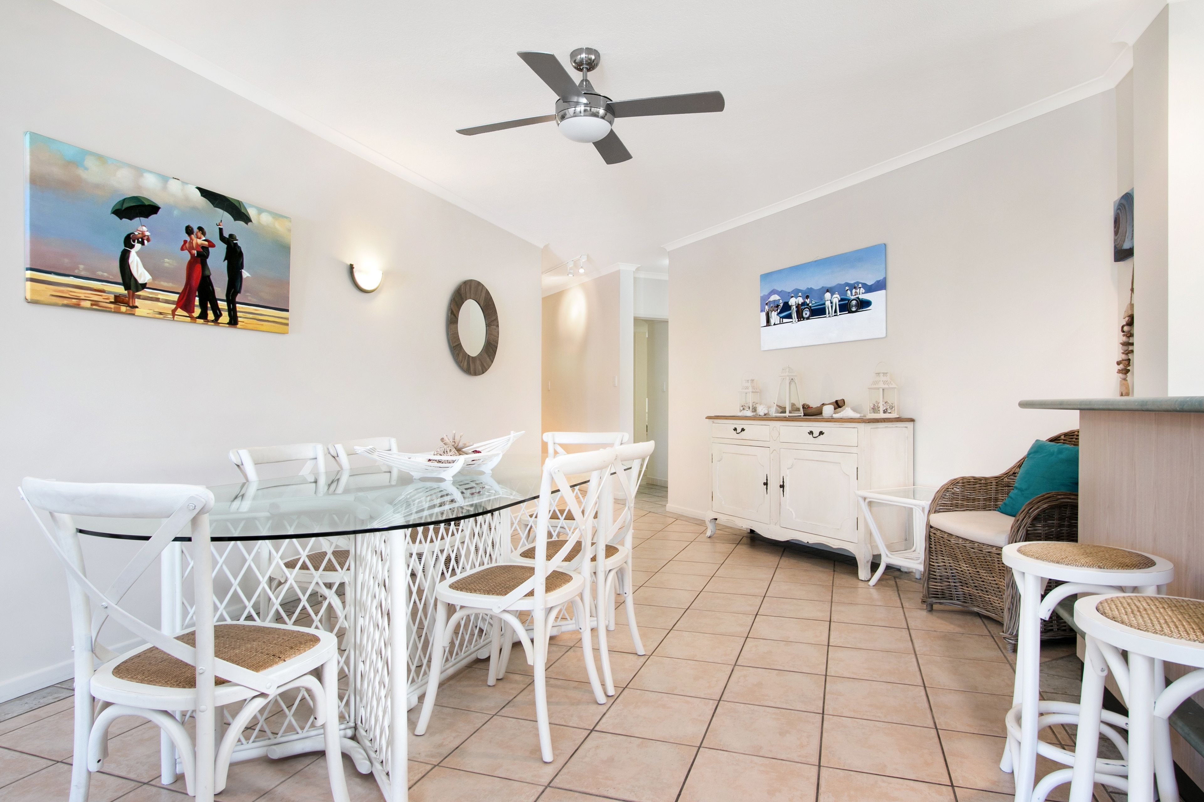 A dining area with a glass table and white chairs, a ceiling fan, and artwork on the walls.