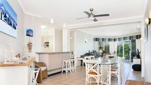 Une cuisine spacieuse avec des meubles blancs, un coin repas doté de chaises en osier et une grande fenêtre offrant une vue sur l’extérieur.