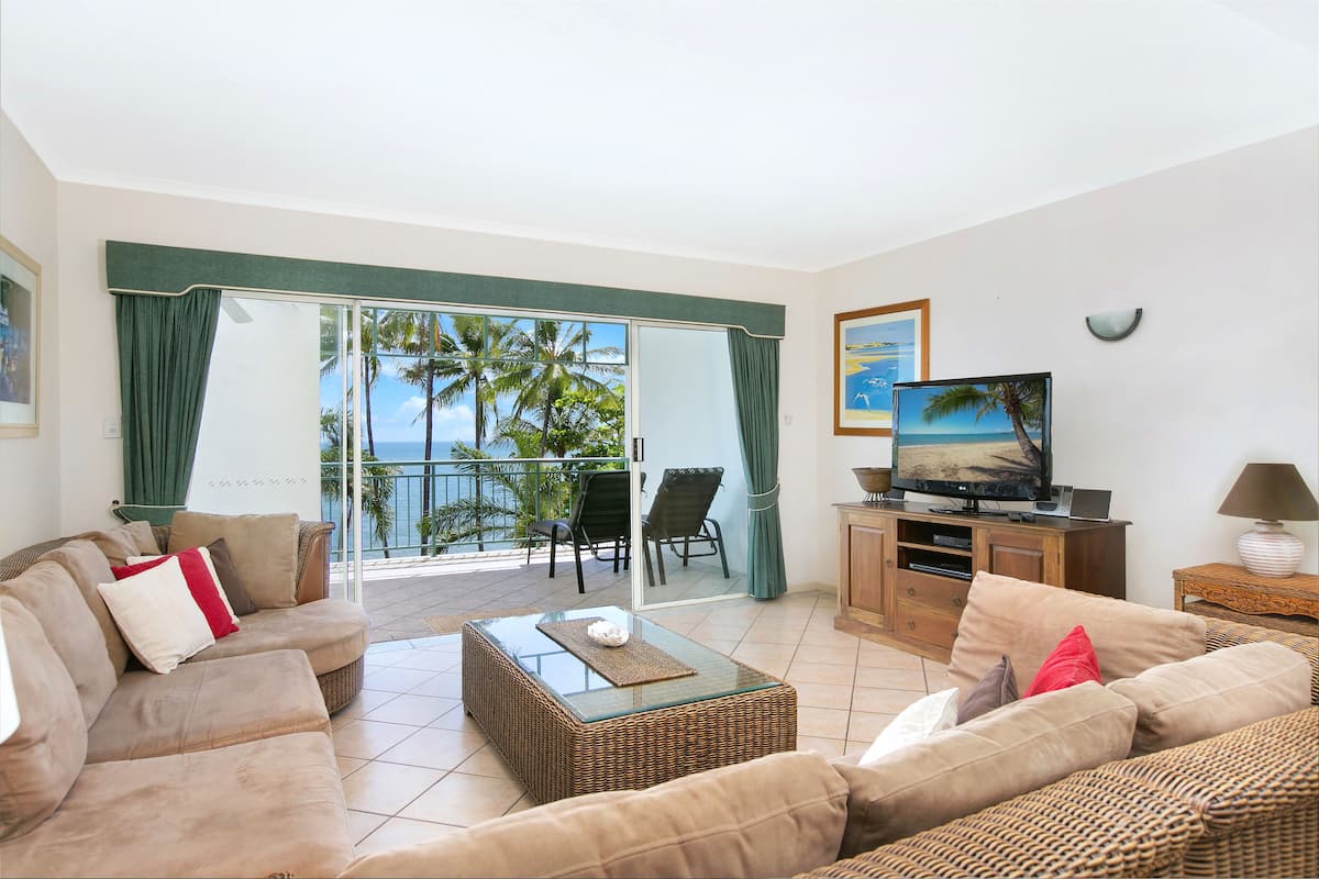 A living room with a sofa, a coffee table, a TV, and a balcony with a view of palm trees and the ocean.