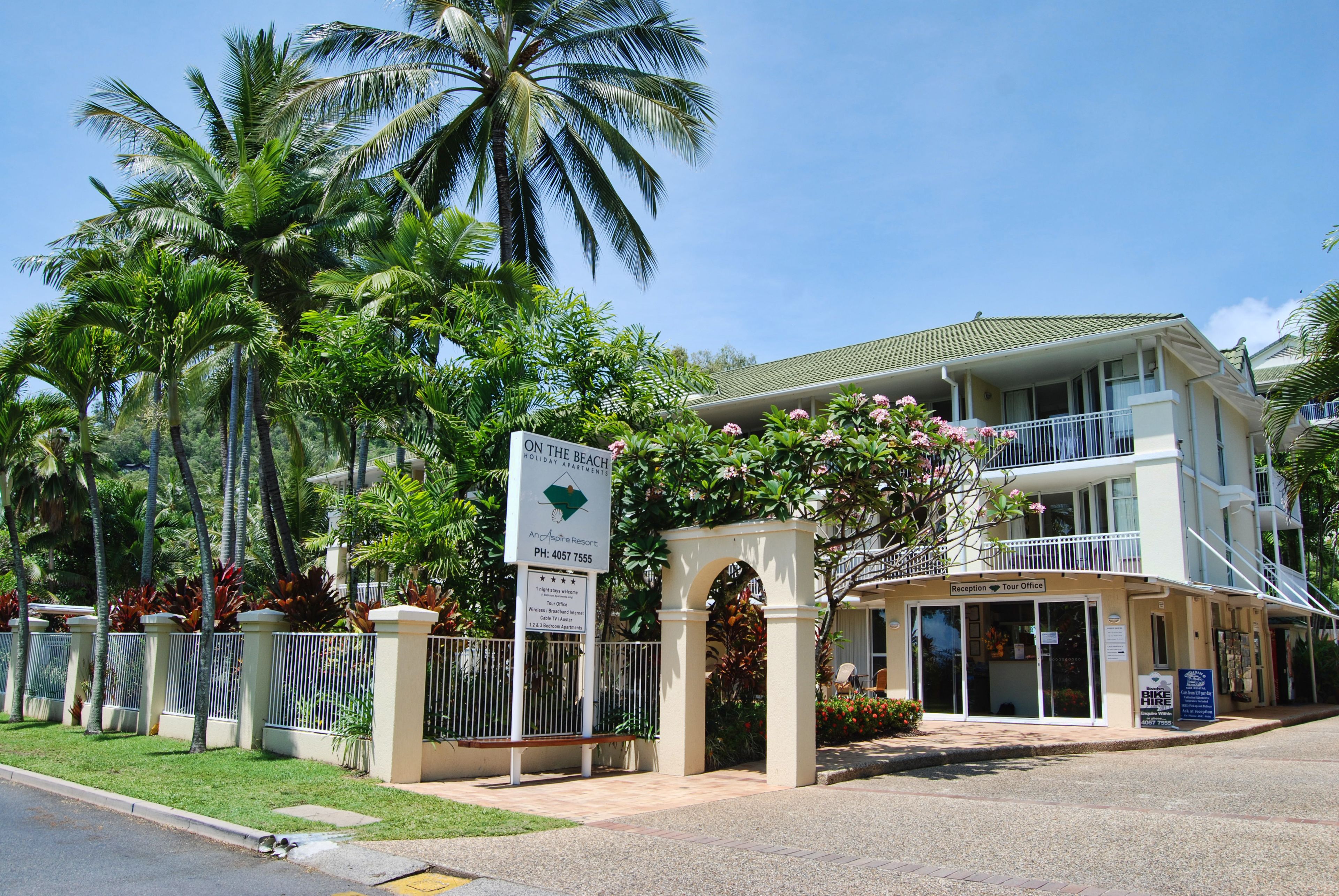 Een hotelentree met een receptiebalie, palmbomen en een bord waarop staat: The Beach Resort.