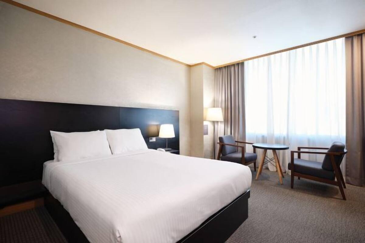 A hotel room with a large bed, a desk, two chairs, a lamp, and a view of the outside through curtains.