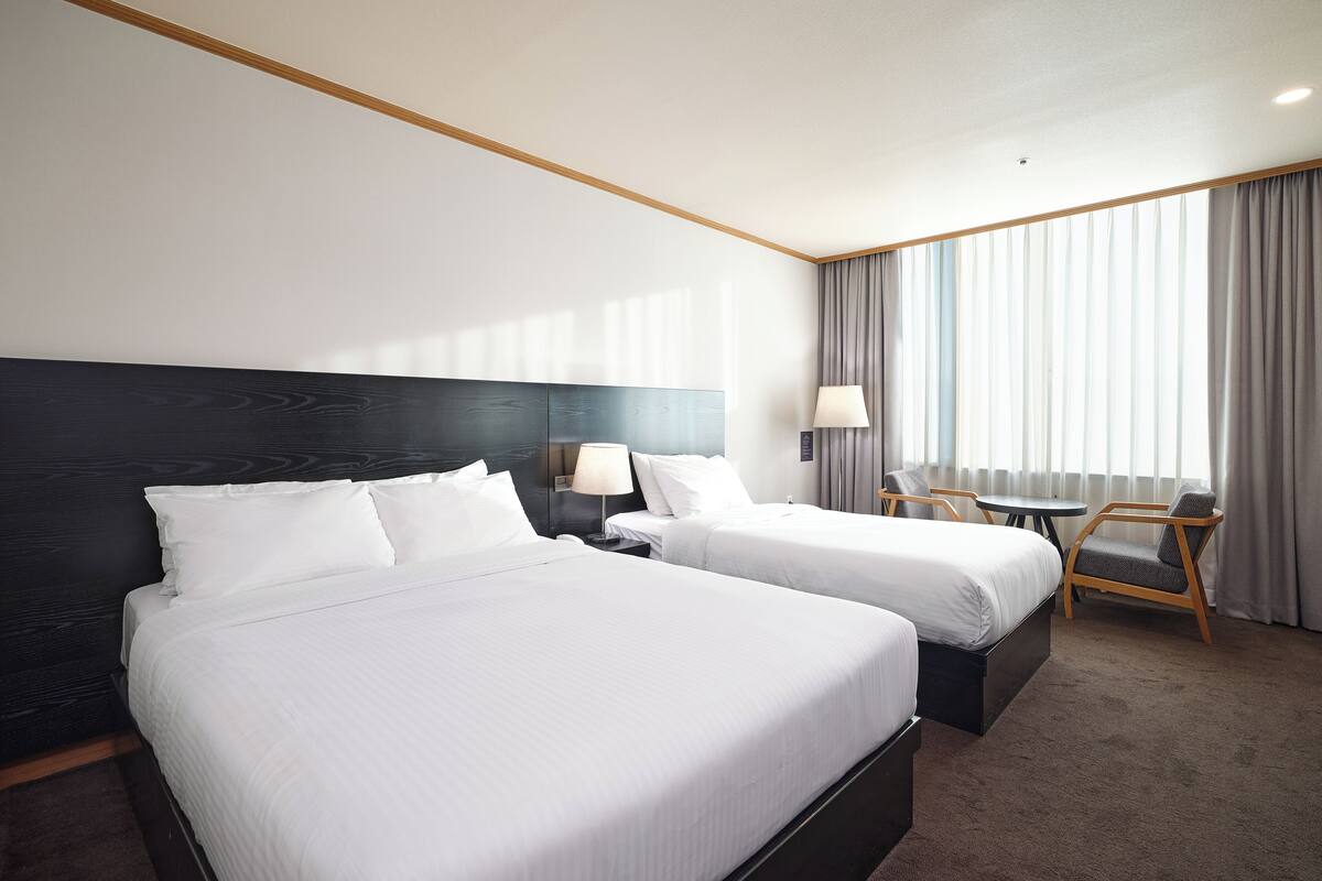 A hotel room with two beds, a desk, a chair, and a large window with curtains.