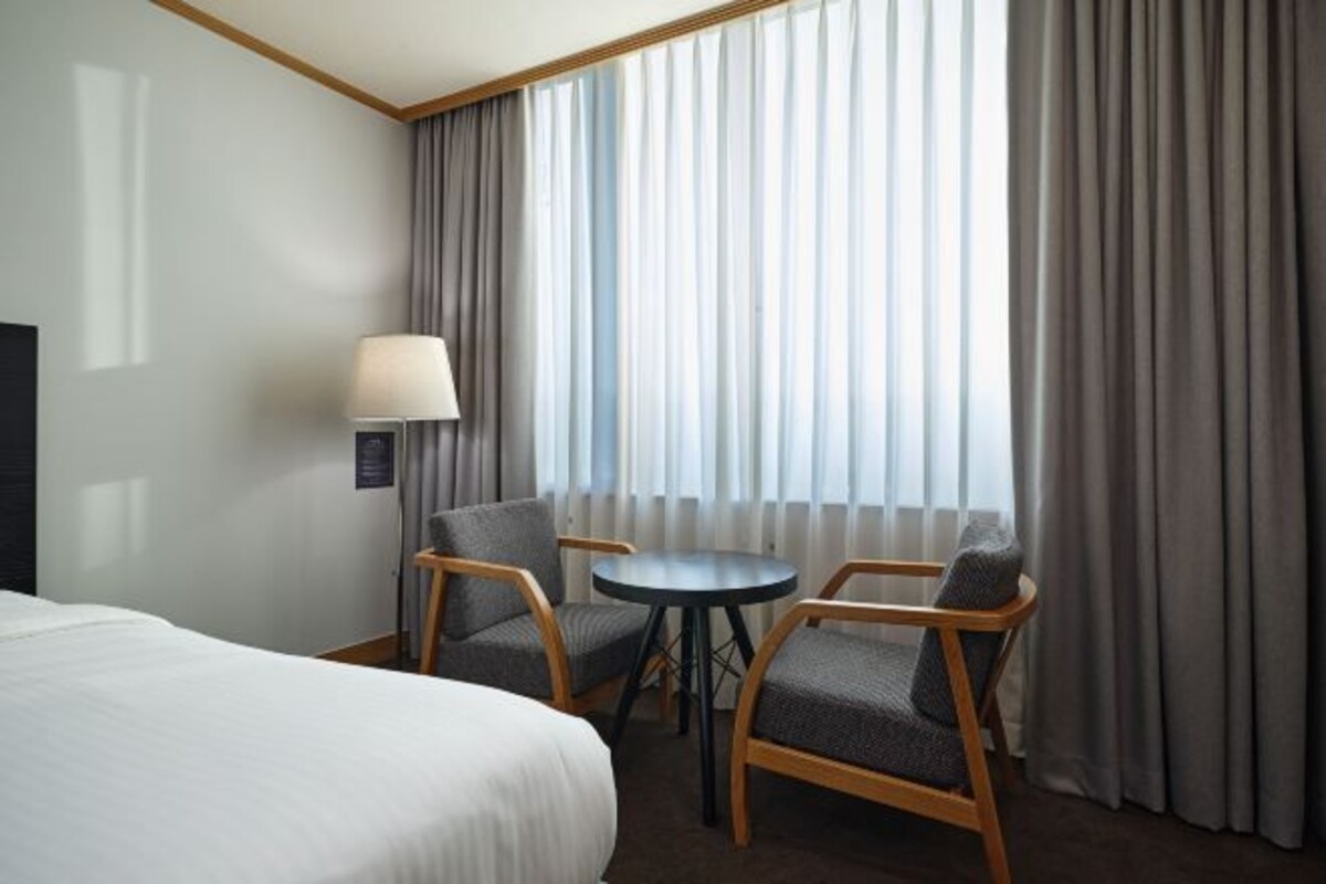 A hotel room with a bed, two armchairs, a small round table, a lamp, and a window with curtains.