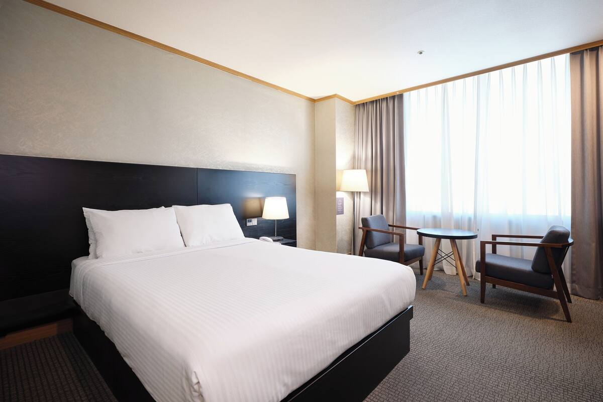 A hotel room with a large bed, a desk with a chair, a lamp, and a window with curtains.