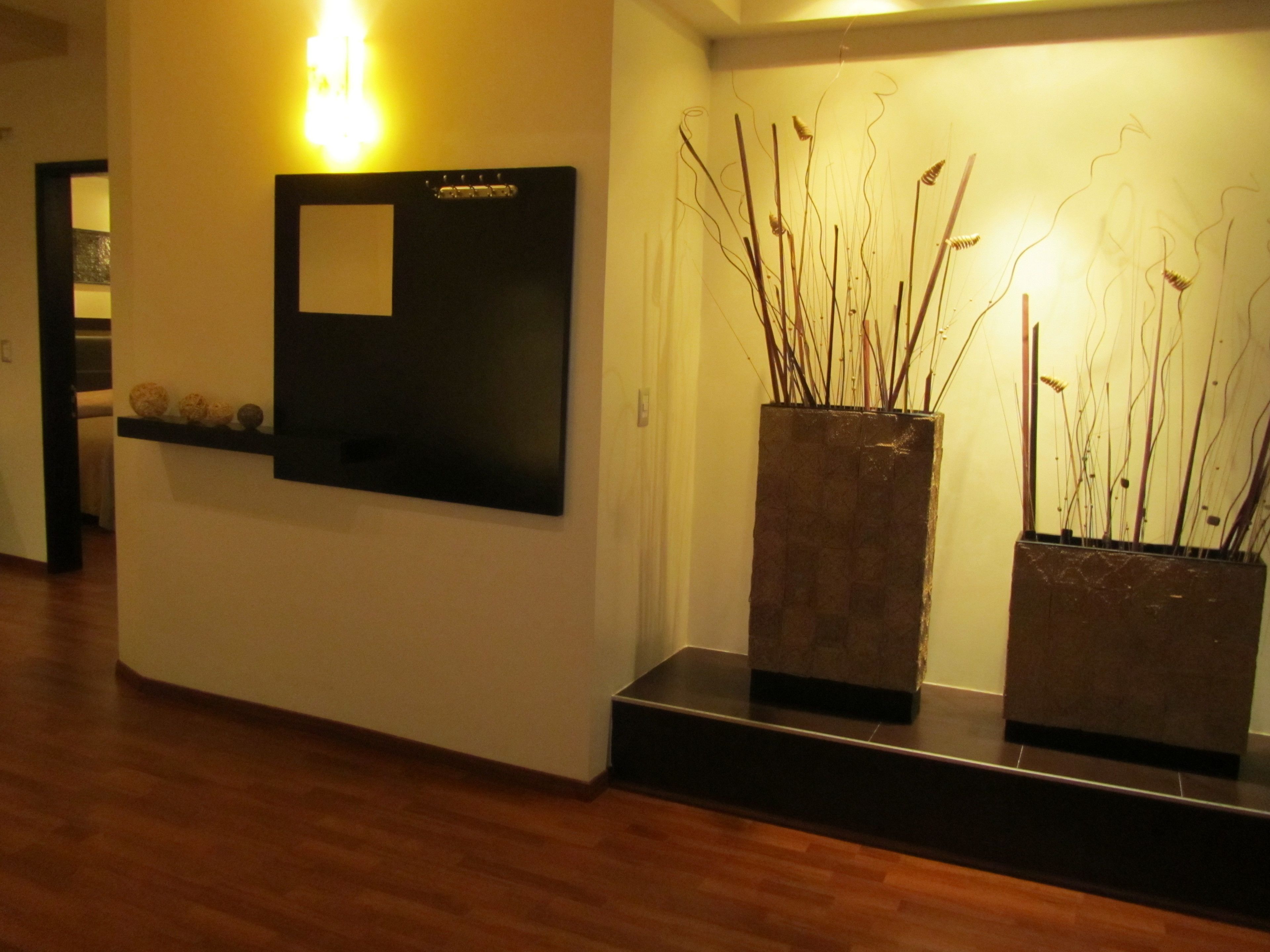 A hallway with a wooden floor, a wall-mounted light fixture, a blackboard, and two planters with tall, thin plants.