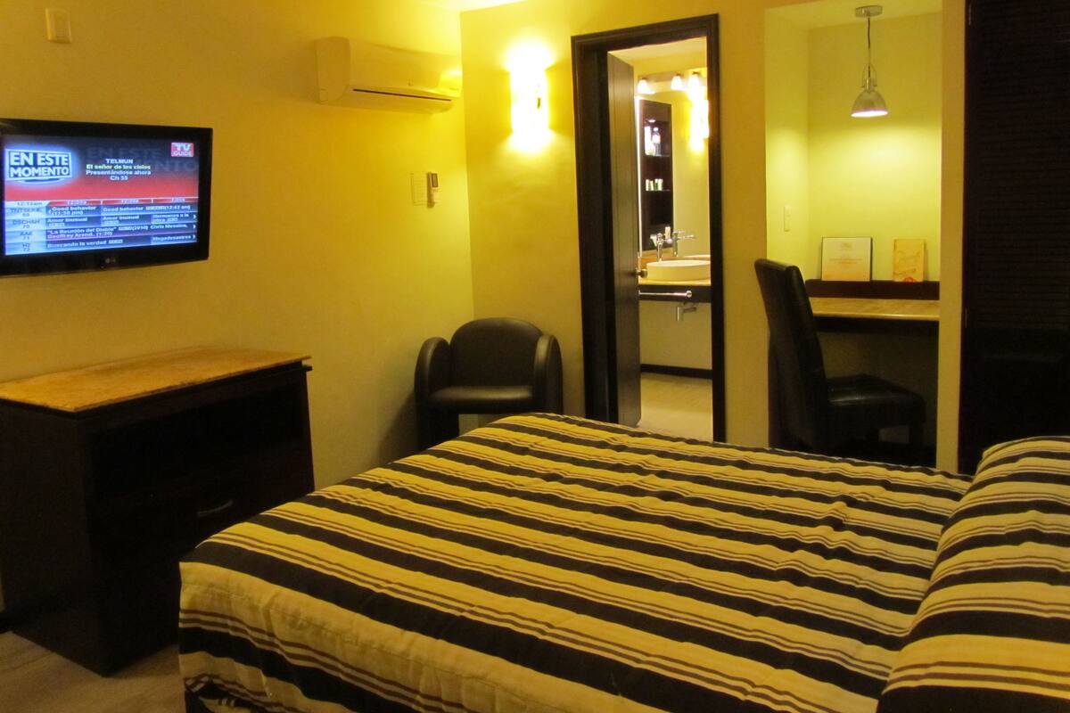 A hotel room with a bed, a desk, a TV, a chair, and a bathroom visible through an open door.