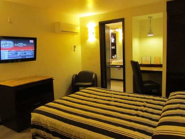 Habitación de hotel con cama, escritorio, televisión, silla y baño visible a través de una puerta abierta.
