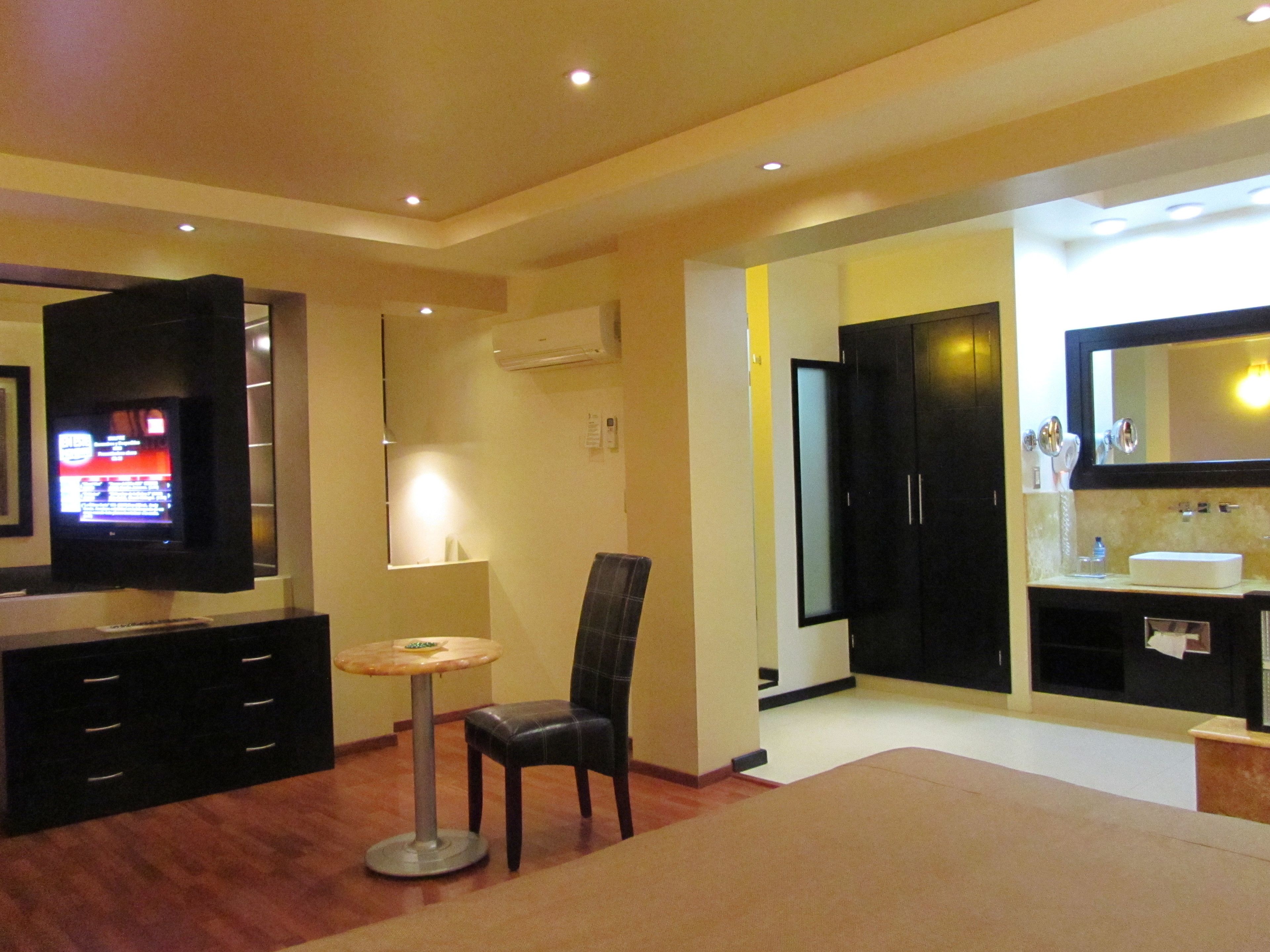 Une chambre d’hôtel équipée d’une télévision, d’une petite table ronde, d’une chaise, d’un lit, d’une armoire et d’une salle de bain avec un miroir et un lavabo.