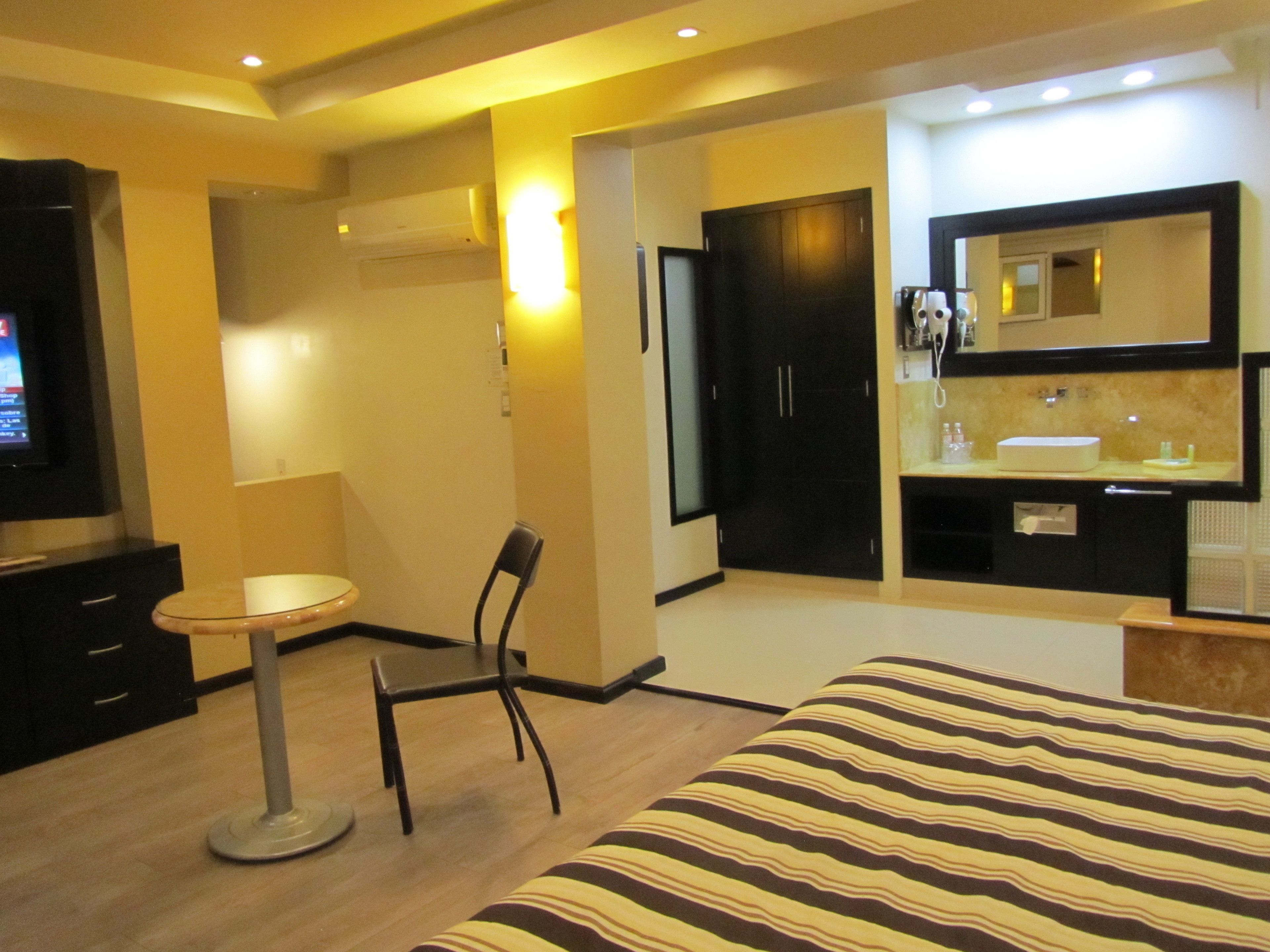 Habitación de hotel con cama, escritorio, silla, televisión y baño con espejo y lavamanos.