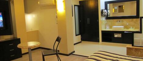 Habitación de hotel con cama, escritorio, silla, televisión y baño con espejo y lavamanos.