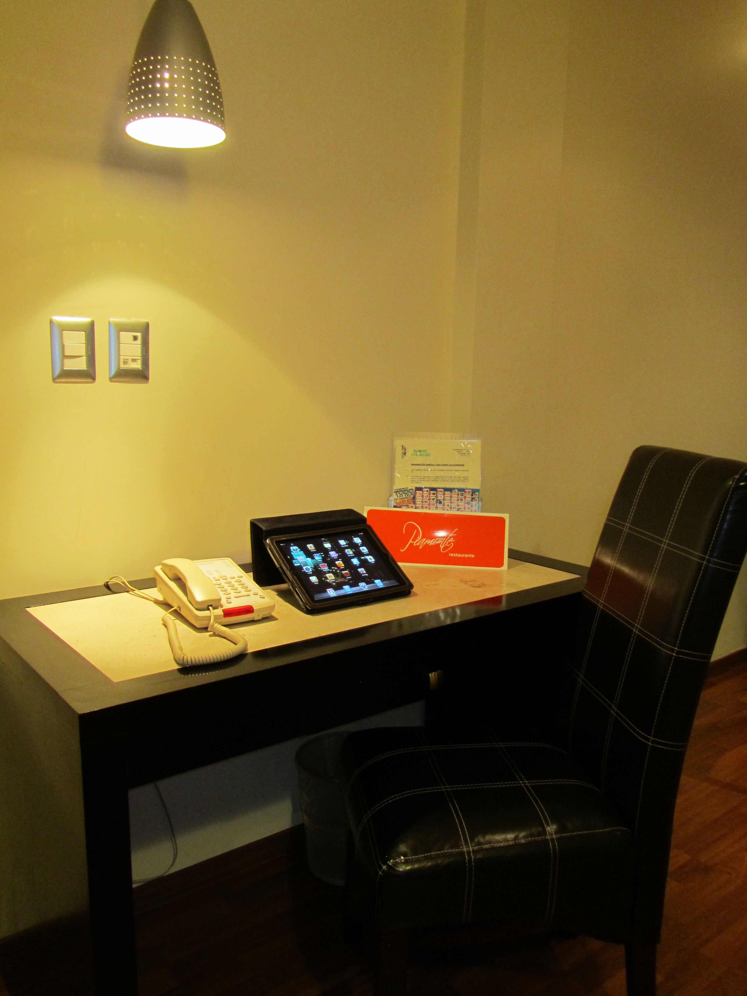 A desk with a phone, tablet, and a red sign, a black chair, and a wall-mounted light fixture.