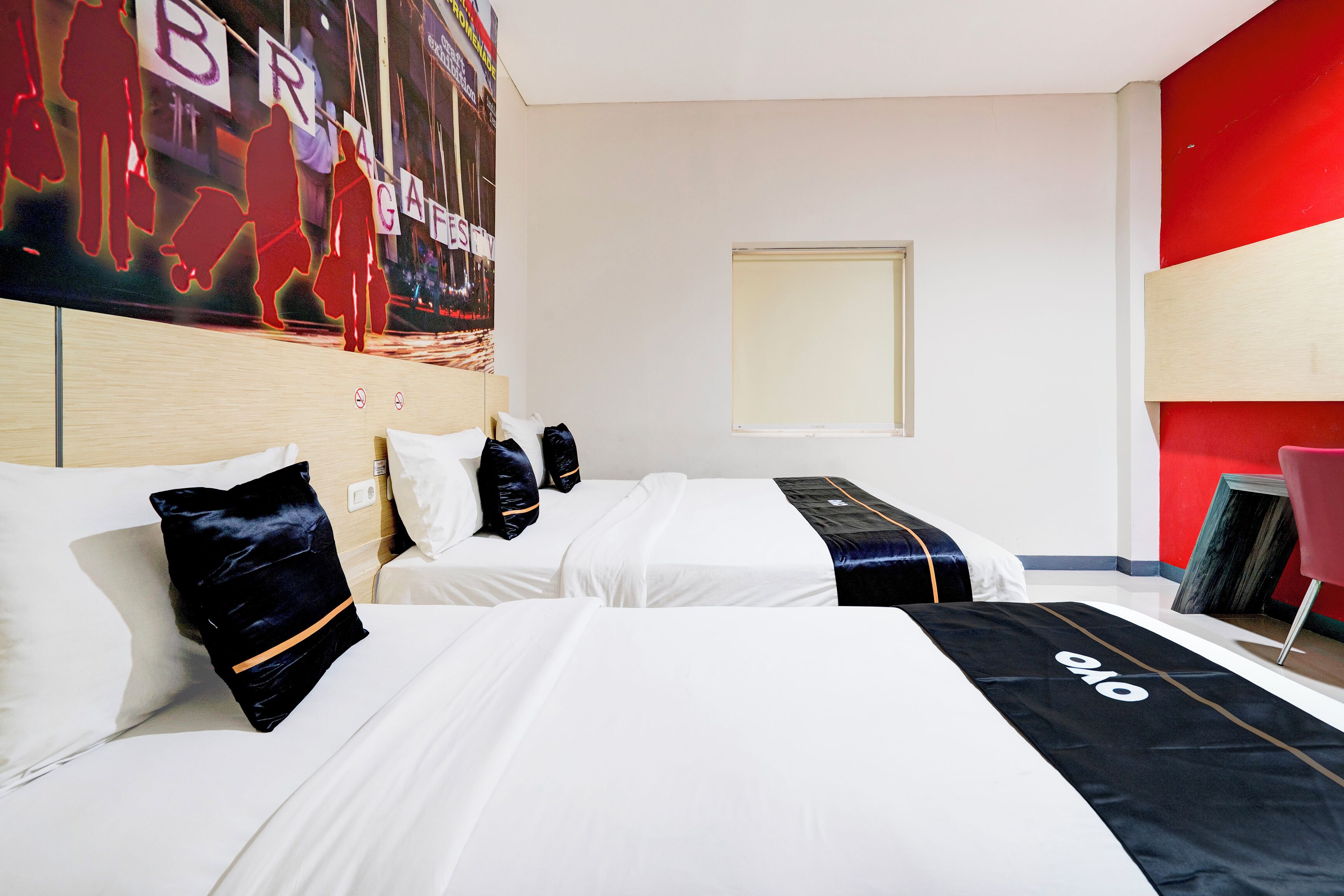 A hotel room with two beds, a red accent wall, a large poster, and a window.