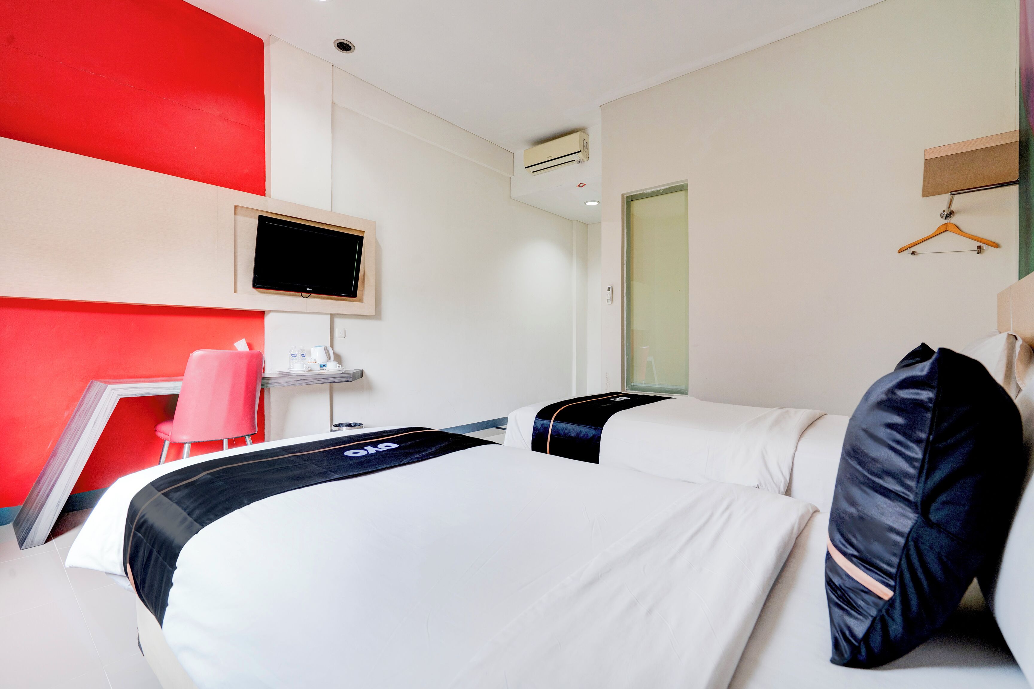 Ein Hotelzimmer mit zwei Betten, einem an der Wand befestigten Fernseher, einem Schreibtisch mit Stuhl und einer roten Wand.