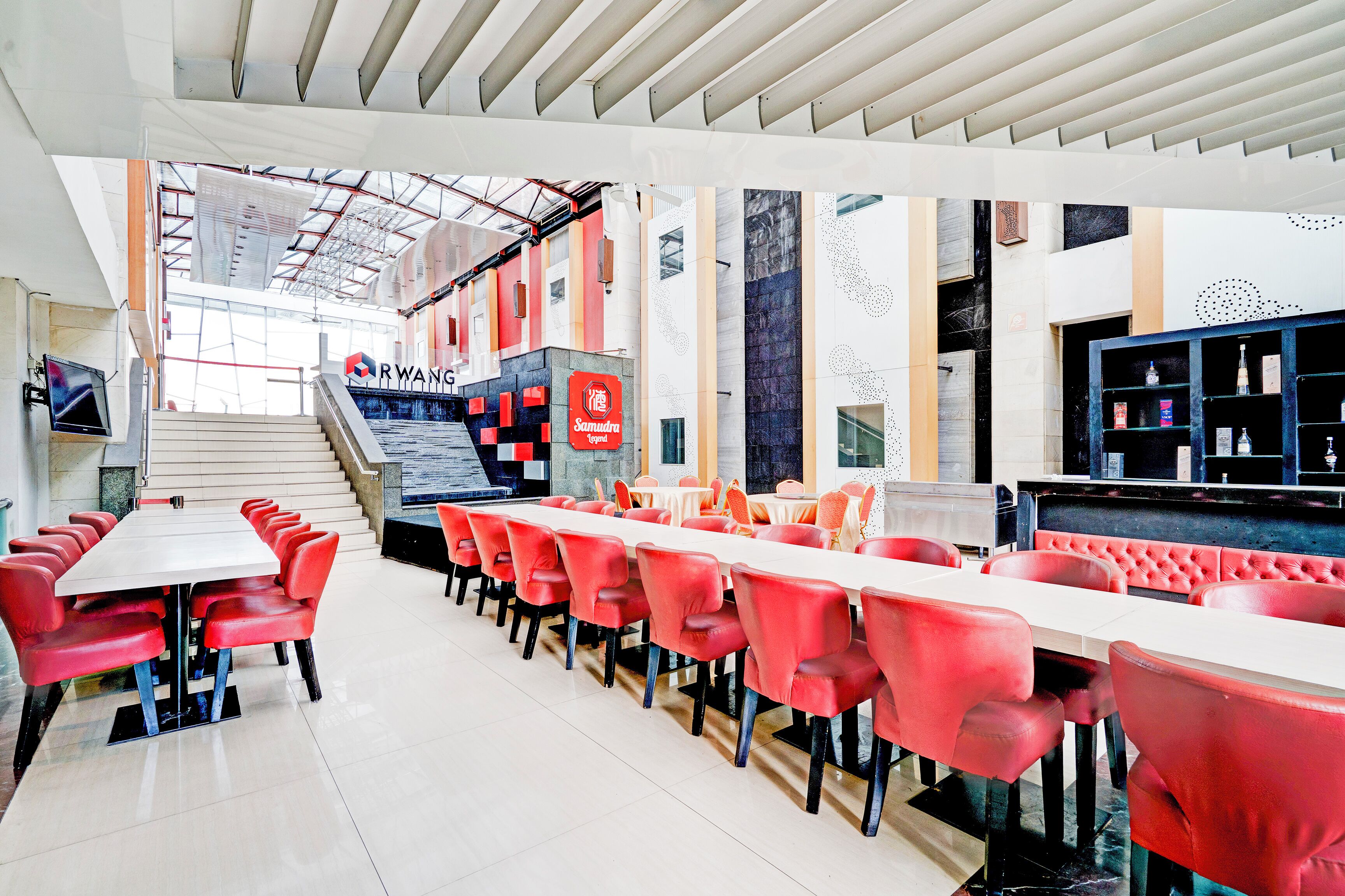 A modern restaurant with red chairs and a long table, a staircase in the background, and a counter with shelves.