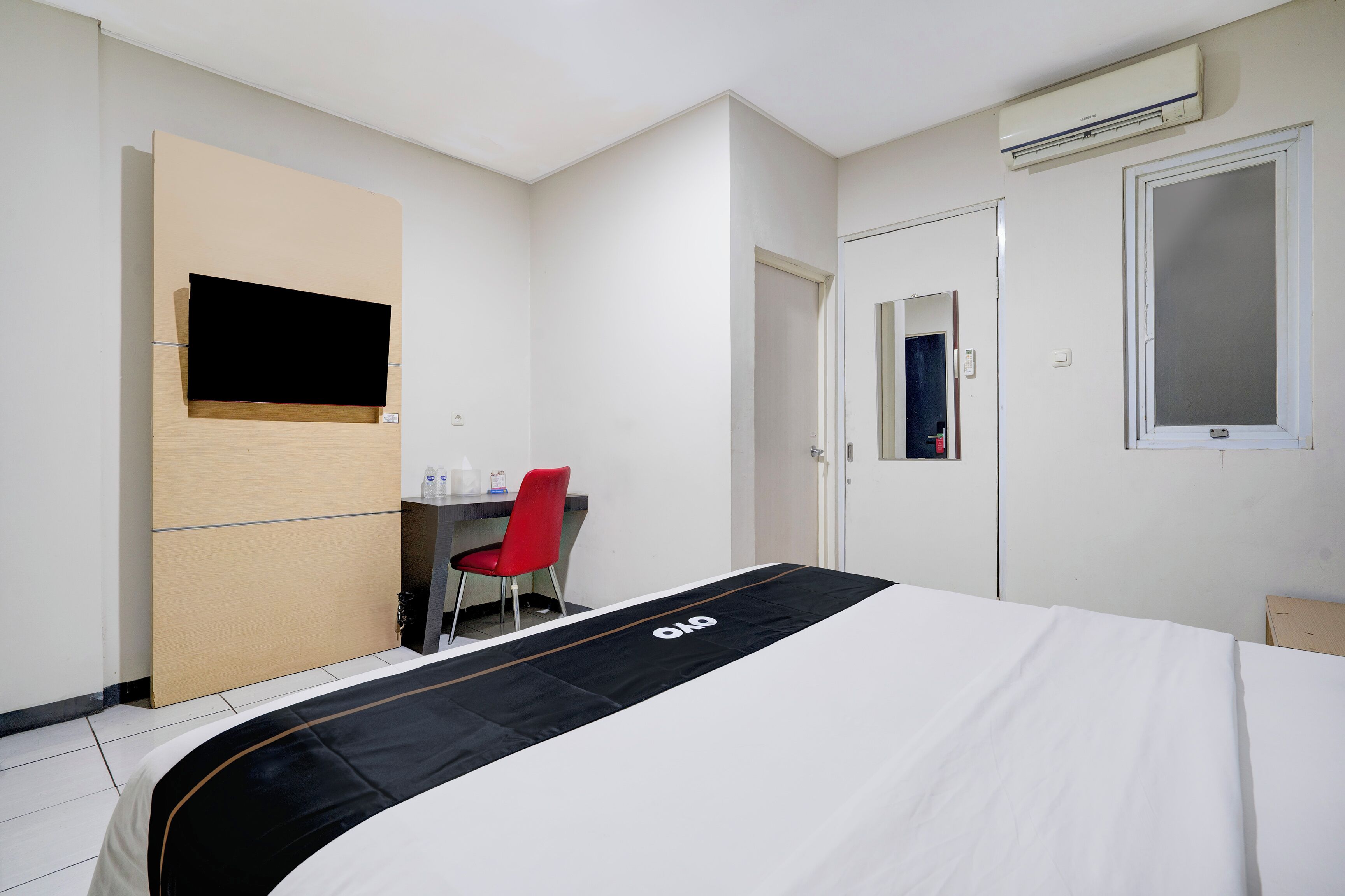 A hotel room with a bed, a desk, a chair, a TV mounted on the wall, and an air conditioning unit.