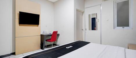A hotel room with a bed, a desk, a chair, a TV mounted on the wall, and an air conditioning unit.