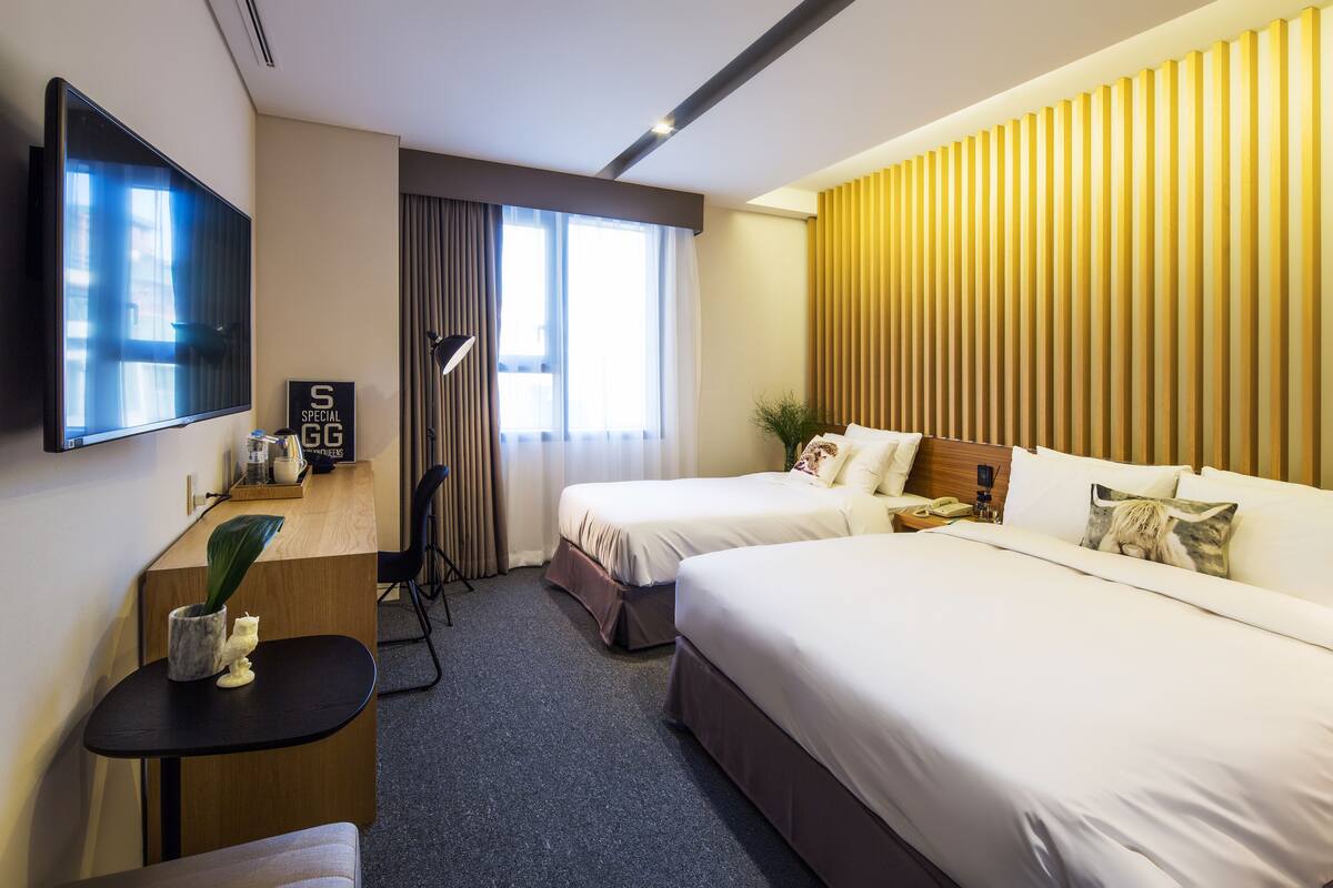 A hotel room with two beds, a desk, a TV, and a large window with curtains.