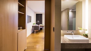 A modern hotel room with a wooden wardrobe, a desk with a chair, a large mirror, and a bathroom with a sink and potted plant. - Hotel Denim (Seoul)