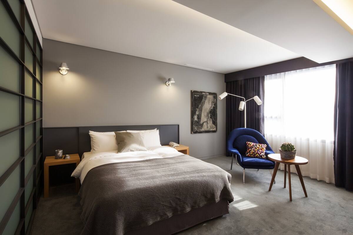 A modern hotel room with a large bed, bedside tables, a desk, a chair, and a window with curtains.