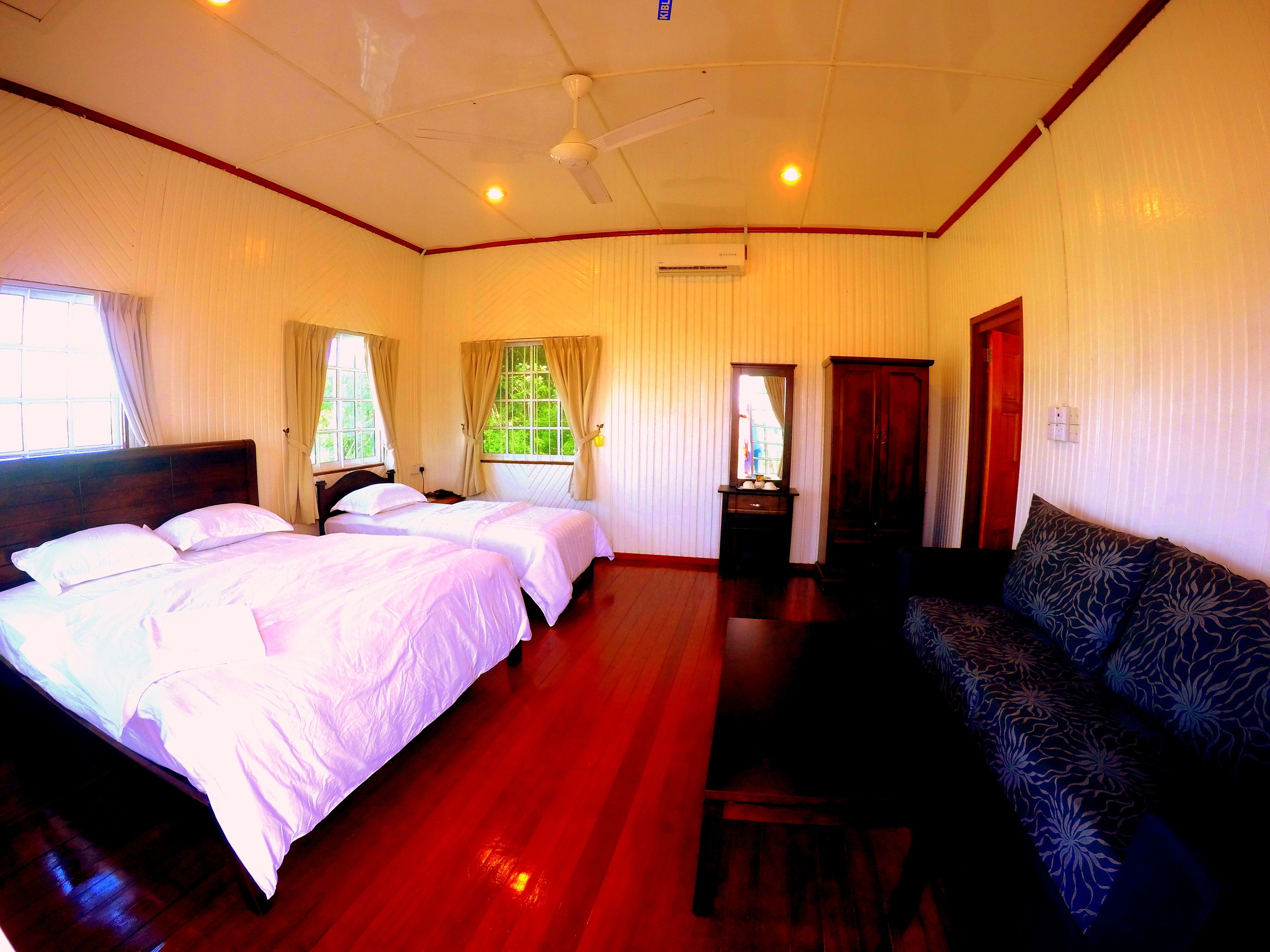A hotel room with two beds, a sofa, a wooden floor, a ceiling fan, and a window with curtains.