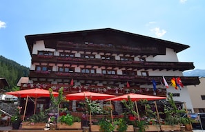 A traditional chalet-style building with multiple balconies adorned with flowers, outdoor seating with red umbrellas, and flags indicating international locations. - Basur - das Schihotel am Arlberg (Flirsch)