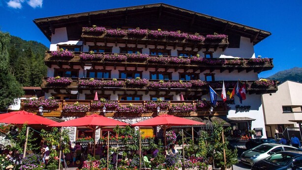 Un hotel de varios pisos con balcones decorados con flores, mobiliario exterior con sombrillas rojas y banderas que denotan reconocimiento internacional.